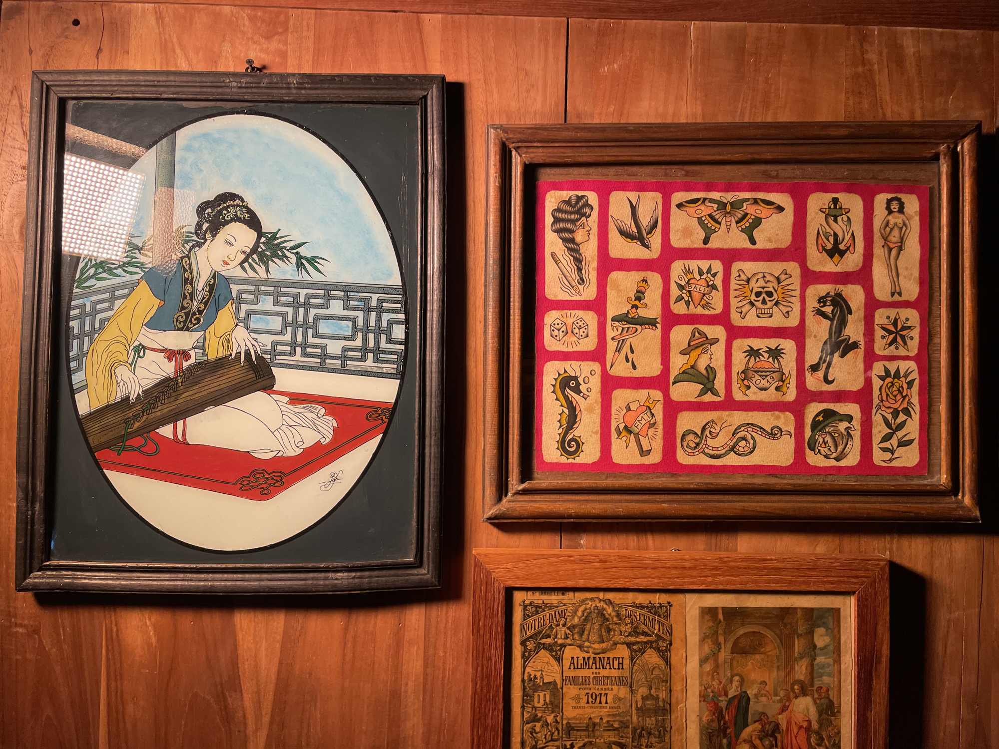 This media file showcases three framed artworks displayed on a warm-toned wooden paneled wall in Mengwi, Indonesia.
On the left, a large, dark-framed painting depicts an East Asian woman in traditional attire, seated and gracefully playing a stringed instrument, possibly a guzheng or koto. The artwork features an oval-shaped inner composition showing the woman against a light blue sky, with a decorative balcony and bamboo foliage. The painting's surface shows reflections, indicating a protective glass or acrylic layer.
To the right, a horizontal, light brown wooden frame encloses a vibrant collection of traditional tattoo flash designs. These small, individual artworks are arranged in a grid on a bright pink background, featuring motifs such as a woman's head, a swallow, a butterfly, an anchor, a pin-up girl, dice, a dagger with a snake, a skull, a panther, a seahorse, a heart with "FMT", a snake, a man in a hat, a tropical scene, and a rose.
Below the tattoo flash, a third, smaller framed piece shows an aged publication. Visible text on its cover includes "NOTRE-DAME ALMANACH DES FAMILLES CHRÉTIENNES POUR L'ANNÉE 1911 TRENTE-CINQUIÈME ANNÉE", along with a number "06341". Adjacent to the almanac cover, another vintage-style religious image is partially visible. The arrangement suggests an eclectic and personal decorative style within an indoor setting.