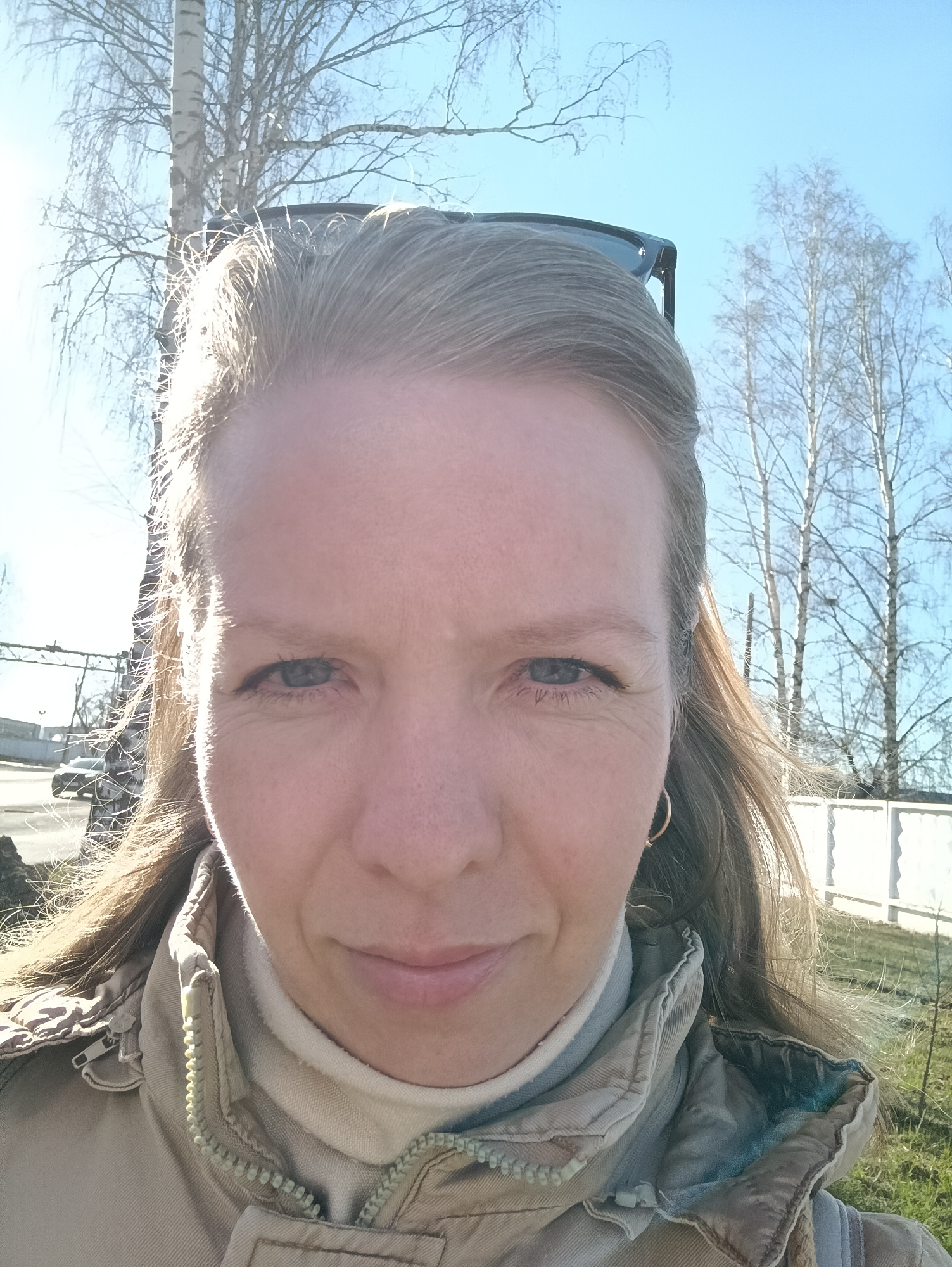 A close-up outdoor selfie depicts a woman, likely in her 30s or 40s, with light brown hair and blue eyes, looking directly at the camera. She wears a neutral-toned, possibly beige or light brown, winter jacket with a visible zipper and a lighter turtleneck underneath. Sunglasses are pushed back on top of her head. Her expression is calm and subtly smiling.
The bright sunlight, casting strong highlights on her hair and the left side of her face, suggests a sunny day, likely morning or afternoon. The background features several bare deciduous trees, characteristic of late autumn, winter, or early spring. One prominent tree, possibly a birch, is visible behind her left shoulder. The sky is clear and blue. To the right, a white fence runs along what appears to be a grassy area, while a road or street with a distant vehicle is visible on the left side, hinting at an urban or suburban setting. The overall scene suggests cool weather. No text is visible in the image. The location context is Chaikovsky, Russia.