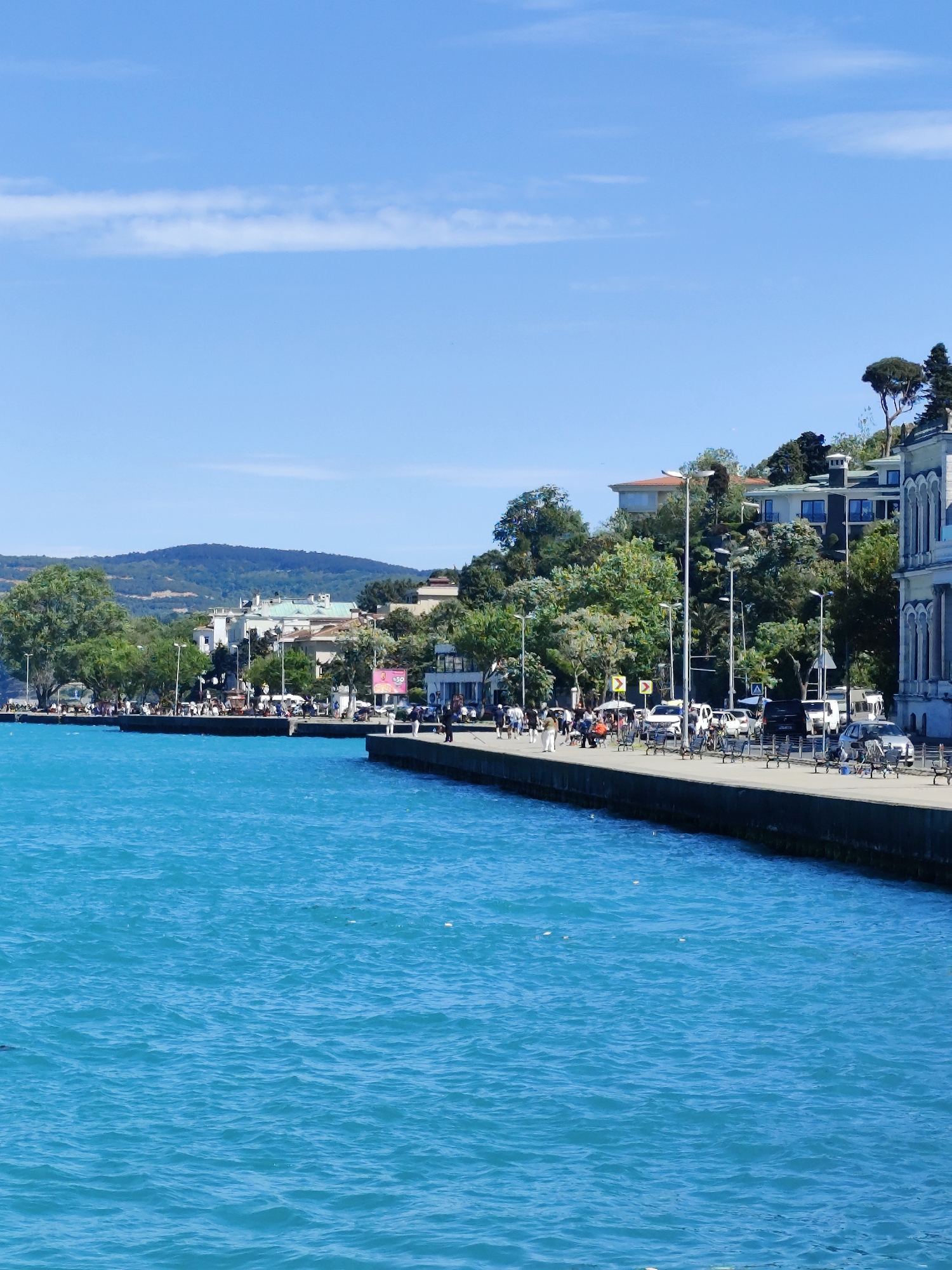 The image captures a scenic view of a waterfront in Sarıyer, Turkey, showcasing a vibrant turquoise body of water bordered by a concrete pier. Along the pier, people are walking, sitting on benches, and engaging in various activities. Cars are parked nearby and streetlights illuminate the path. The background reveals houses nestled among lush greenery on a hill, with a distant mountain range adding depth to the scene. The sky is clear and blue, with a thin white cloud stretching across the horizon.