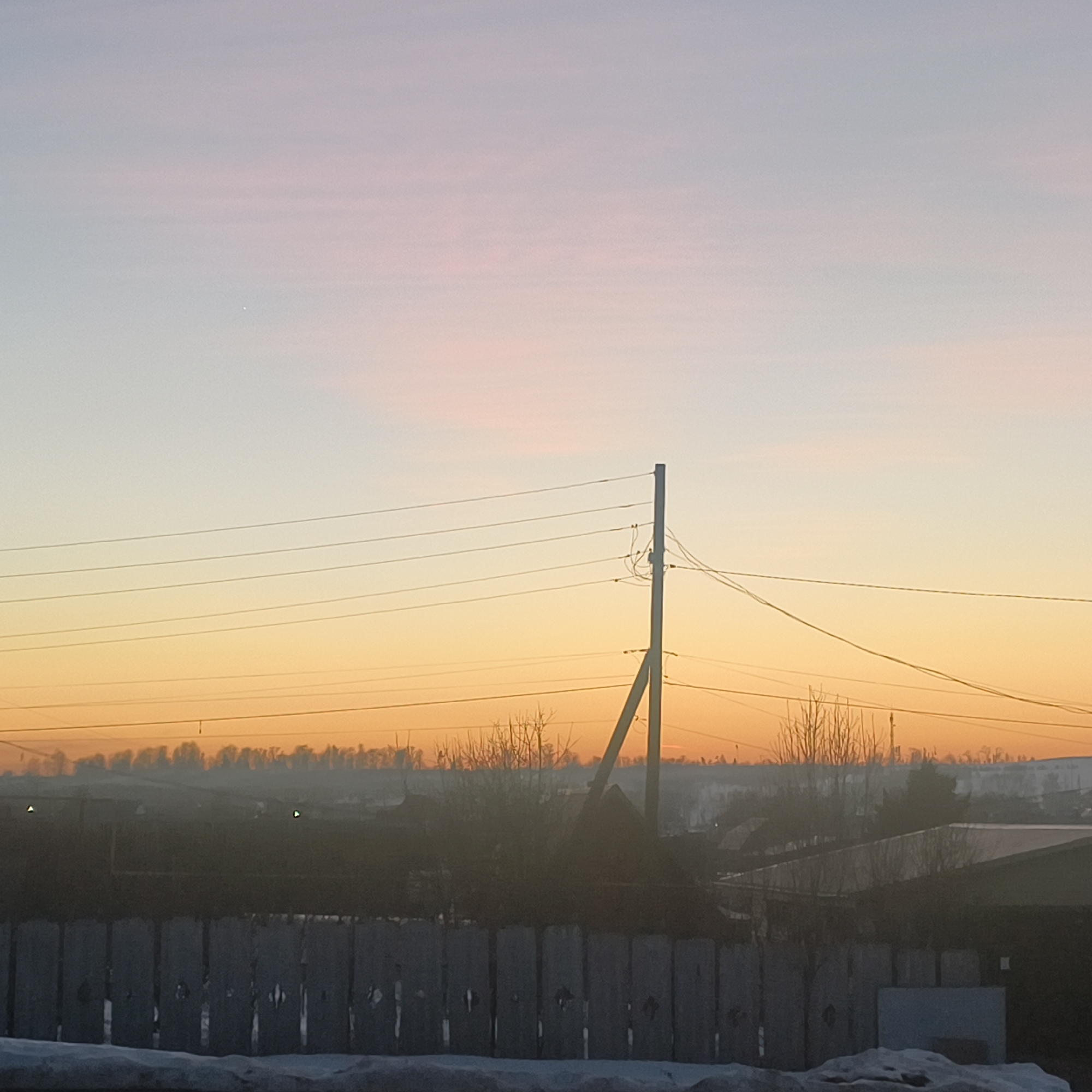 The image depicts an early morning scene in Фоки, Russia. A wooden fence is in the foreground, partially covered in snow. Beyond the fence is a residential area with houses and bare trees. A utility pole with wires is visible in the middle ground. The sky transitions from pink at the top to orange near the horizon. A small, bright point of light is visible in the upper left corner of the sky. A layer of haze obscures the distant landscape.