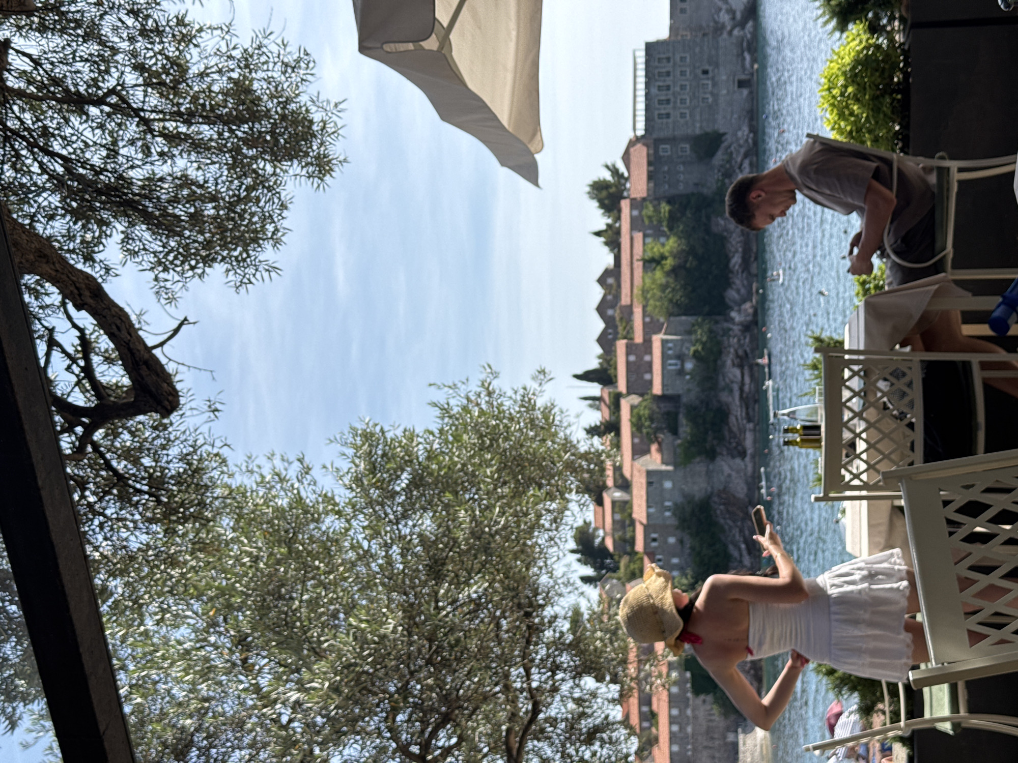 The image captures an outdoor dining scene overlooking the historic fortified island of Sveti Stefan in Montenegro on a clear, sunny day. In the foreground, a woman stands with her back to the viewer, dressed in a white strapless top, a white pleated skirt, and a straw hat with a red ribbon, holding a mobile phone in her right hand with her left hand on her hip. Adjacent to her on the right, a man in a grey t-shirt and dark trousers is seated at a table, looking down at a mobile device held in both hands. The tables are set with bottles and accompanied by white chairs with lattice backs. A large olive tree with dense green foliage occupies the left and upper left portions of the foreground, while dark tree branches extend across the top. A partial white patio umbrella is visible in the upper right. The midground features the blue Adriatic Sea, with small white objects (possibly boats) visible in the distance. Beyond the water, the island of Sveti Stefan is prominent, characterized by numerous multi-story stone buildings with reddish-orange tiled roofs amidst green foliage. The background is a bright, clear blue sky.