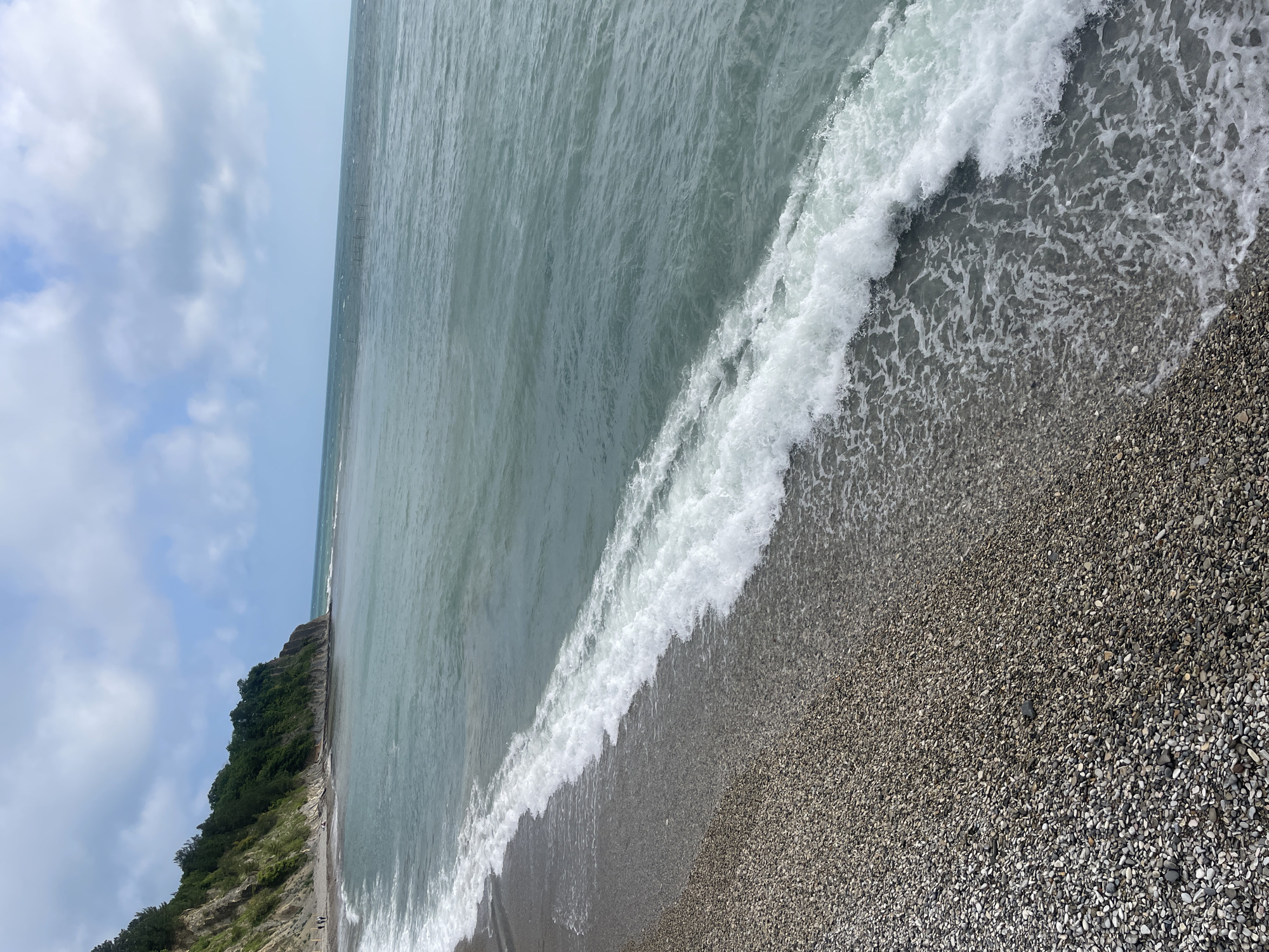 The image shows a beach scene in Olginka, Russia. The beach is covered in small, grey pebbles. Waves with white foam are crashing onto the shore. The sea is a greyish-green color. To the left, there is a coastline covered in green vegetation. The sky is partly cloudy.