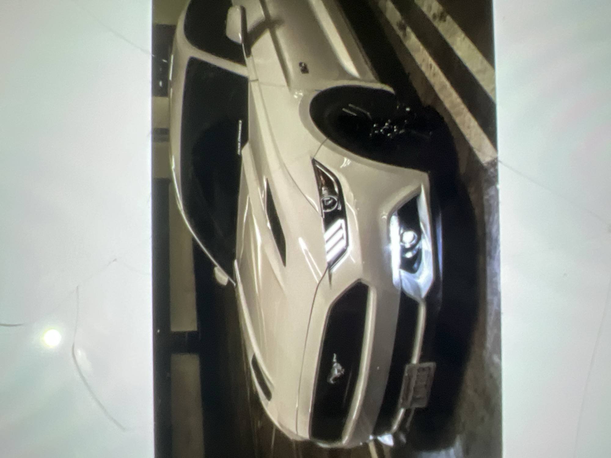 The image shows a white car, possibly a Ford Mustang based on the emblem visible on the grille. The car is parked in what appears to be a parking garage or a similar indoor setting, indicated by the striped markings on the floor which are typical of parking spaces. The lighting suggests it might be nighttime or an indoor environment with artificial lighting, as there are bright spots reflecting on the car's surface. The car is clean and appears to be in good condition. The perspective is from above and slightly to the side, giving a clear view of the car's hood, headlights, and front grille. There is no visible text on the car itself, but there might be some indistinct markings on the wall or floor in the background. No people are visible in the image.