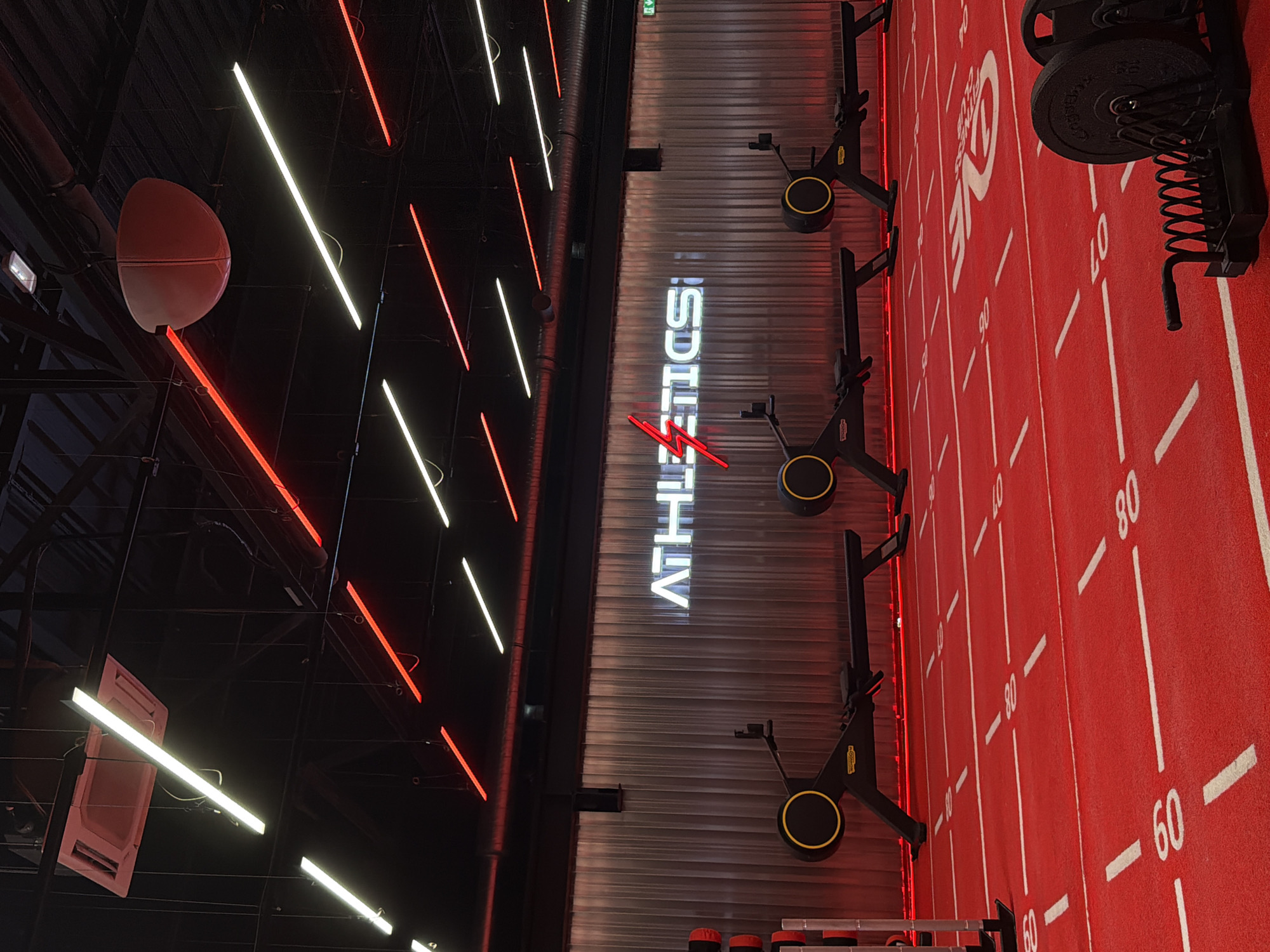 The image depicts the interior of a modern gym, likely located in Strasbourg, France. The gym features a vibrant red floor with white markings indicating distances or zones. In the background, a corrugated metal wall serves as a backdrop for a neon sign that reads "ATHLETICS!" in white lettering with a red lightning bolt graphic. Several rowing machines, identifiable by their black frames, yellow accents, and the "Technogym" logo, are lined up in front of the wall. The ceiling is a dark industrial style, with exposed beams and a network of lighting fixtures, including white and red LED strips and a large, pink, dome-shaped speaker or light fixture. The overall impression is one of a high-energy, well-equipped fitness facility. There are no people visible in the image.