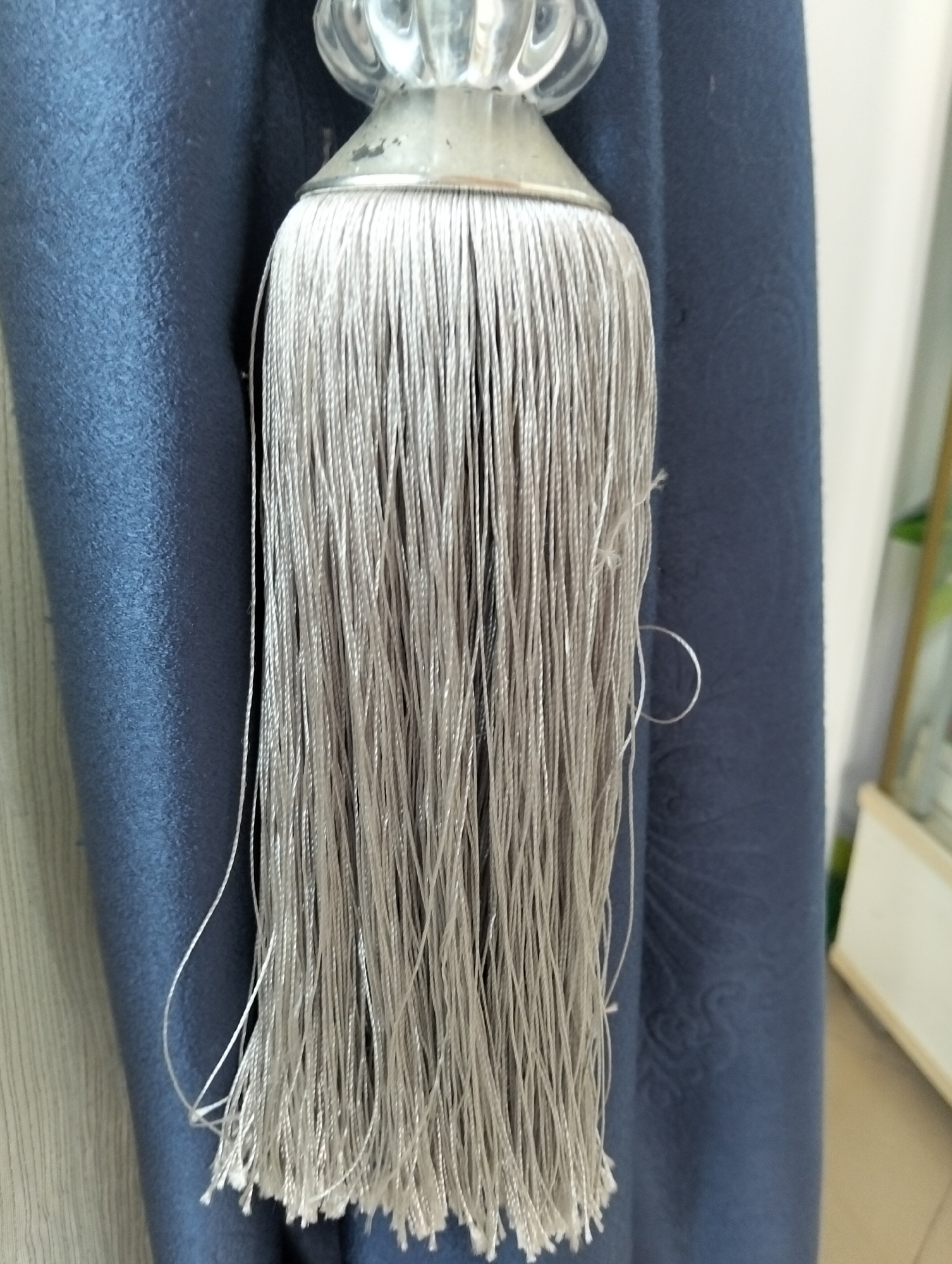 A close-up, eye-level, medium shot captures a silver tassel hanging from a dark blue curtain. The tassel, composed of numerous thin, intertwined threads, hangs downwards, some threads appearing slightly frayed or unraveled at the bottom. Above the tassel, a decorative, clear acrylic finial with an ornate, faceted design is visible, attached to a metallic cap from which the tassel's threads originate. The blue curtain to the right of the tassel has a subtle, embossed pattern. To the left of the tassel, another curtain or fabric surface is visible, appearing to be a lighter, textured material with vertical lines. The background suggests an indoor setting, with a portion of a white cabinet or shelf visible in the upper right corner, and the floor or a surface in the lower right corner, suggesting a domestic environment. There are no people present in the image. The lighting appears to be natural, casting soft shadows. The location is in the city of Julicotmeurak, Indonesia.