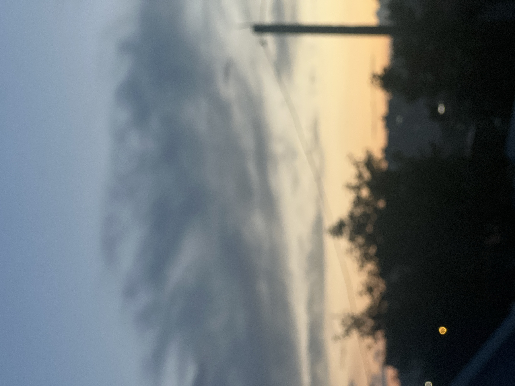 The image depicts a blurred view of a city skyline at dusk. In the foreground, silhouetted trees and a utility pole are visible against a gradient of sky colors, ranging from a light blue at the top to a warm orange near the horizon. A thin power line stretches across the frame. In the background, indistinct shapes suggest buildings and other urban elements. Two bright, circular bokeh lights are present in the lower portion of the image, one appearing yellow and the other white. The overall impression is that of a slightly overcast evening in an urban environment, potentially Almetyevsk, Russia, given the prompt.