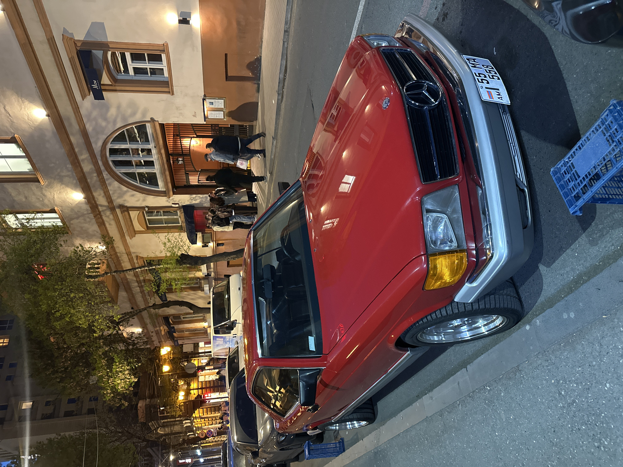 The image captures a red Mercedes-Benz parked on a street in Yerevan, Armenia. The license plate is visible, reading "I 55 MA 558" with "AM" below. A blue plastic crate sits beside the car. Pedestrians are seen entering a building behind the car, likely a restaurant or establishment called "Mov," judging by the sign above the door. The building features arched windows and warm lighting. Streetlights illuminate the scene, casting shadows and highlighting the architectural details of the surrounding buildings. Other vehicles are parked down the street. The overall atmosphere suggests an evening setting in a lively part of Yerevan.