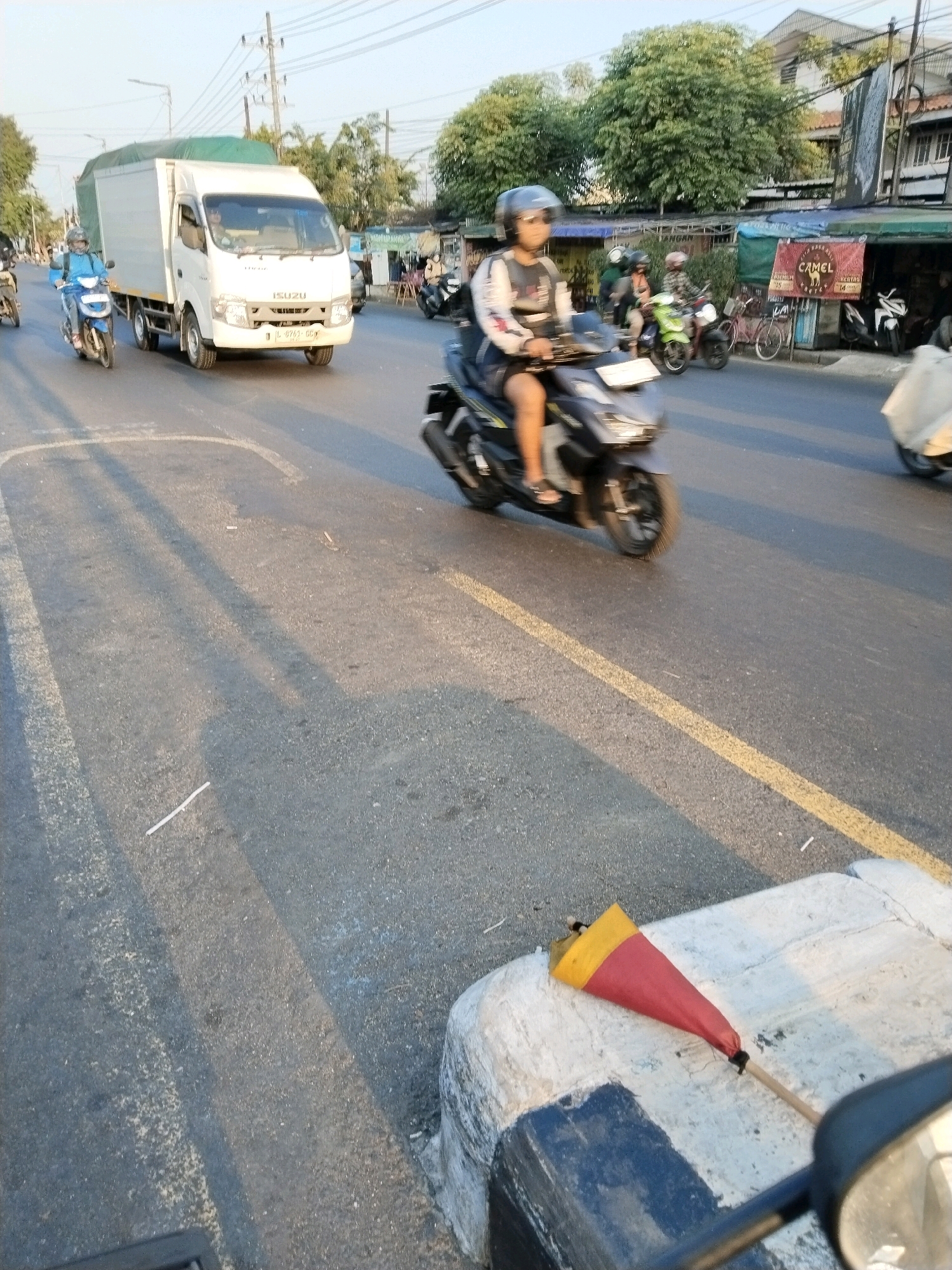 A convoy of vehicles, including motorcycles and a white box truck, proceeds down a street in Medaeng, Indonesia. The truck, bearing the ISUZU logo and license plate L 8763 CC, is prominently positioned. A person on a blue motorcycle rides alongside the truck, while another individual on a dark-colored scooter navigates the road ahead. The street is paved and marked with yellow lines. Along the side of the road, shops with various signs, including one advertising "CAMEL," are visible. Trees line the background, and power lines crisscross the sky. In the foreground, a red and yellow cone-shaped object rests on a concrete barrier, with a motorcycle mirror partially obscuring the view. The scene is bathed in the warm light of a low-hanging sun, casting long shadows across the asphalt.