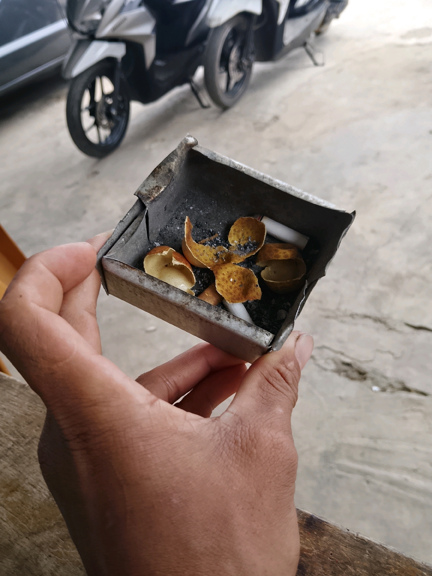 A person's hand holds a small, square metal ashtray containing cigarette butts and what appear to be dried fruit peels. The ashtray is worn and appears to be made of tin. The background is blurred and shows a portion of a motorcycle and a concrete ground. The overall scene is outdoors in an unspecified urban or semi-urban location.