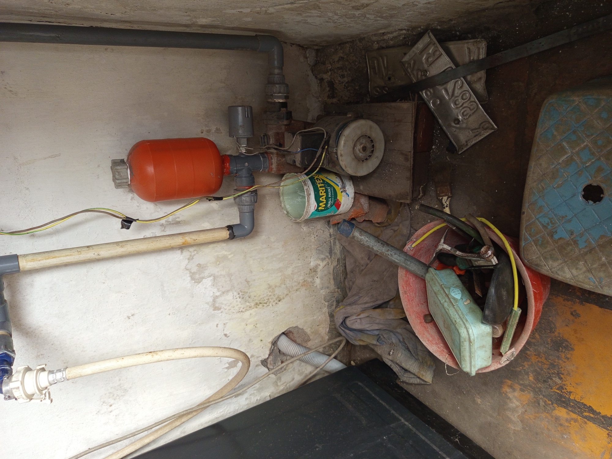 The image depicts a cluttered utility area, possibly in Tebel, Indonesia. In the upper left, a white hose connects to a blue fitting and then to a PVC pipe. A wooden pole runs vertically on the right of this pipe. In the center, an orange pressure tank is mounted on a pipe system, which connects to a rusty water pump with visible wiring. A green and white paint bucket labeled "MARITEX WALL PAINT" sits next to the pump. Below the pump, a wooden board supports the equipment, with a silver fan to its right. In the foreground, a red plastic basin overflows with various items, including a light blue container with a lid, tools, and a yellow hose. Rags are piled to the left of the basin. To the right of the basin, two metal plates with embossed lettering and numbers, resembling license plates, are stacked. A blue plastic stool with a circular opening is partially visible in the bottom right corner. The walls appear weathered and stained, and the concrete floor is uneven and discolored with yellow markings. The overall impression is one of disarray and disuse.