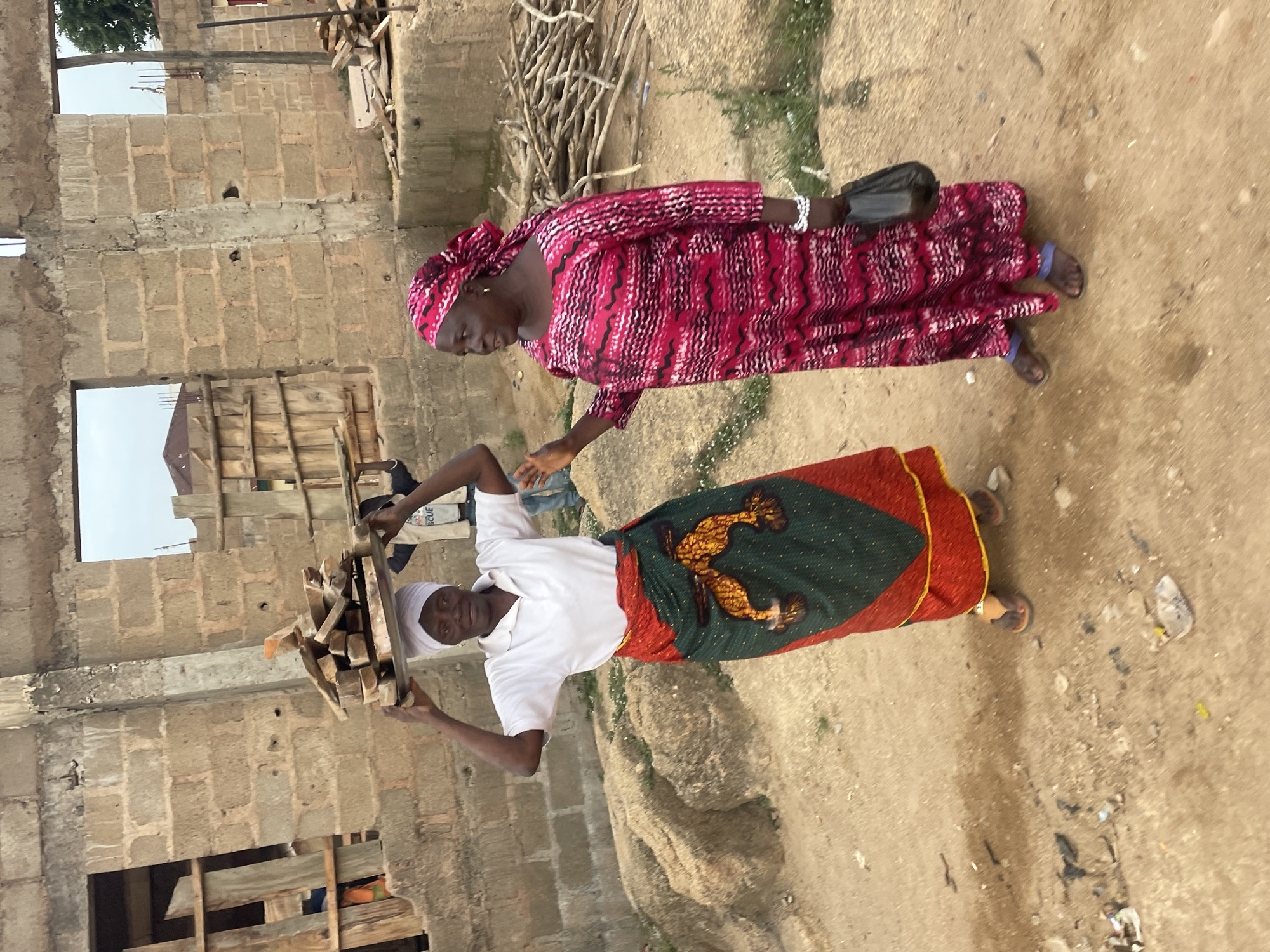 Two women are outdoors in Jos, Nigeria. One woman, wearing a white top, a colorful patterned skirt, and a white head covering, carries a bundle of wood on her head. She is smiling. The second woman, wearing a long red and pink patterned dress and matching head covering, walks beside her. The environment includes a partially constructed building made of cement blocks, wood planks, and some other wood piles behind them. The ground is dirt.