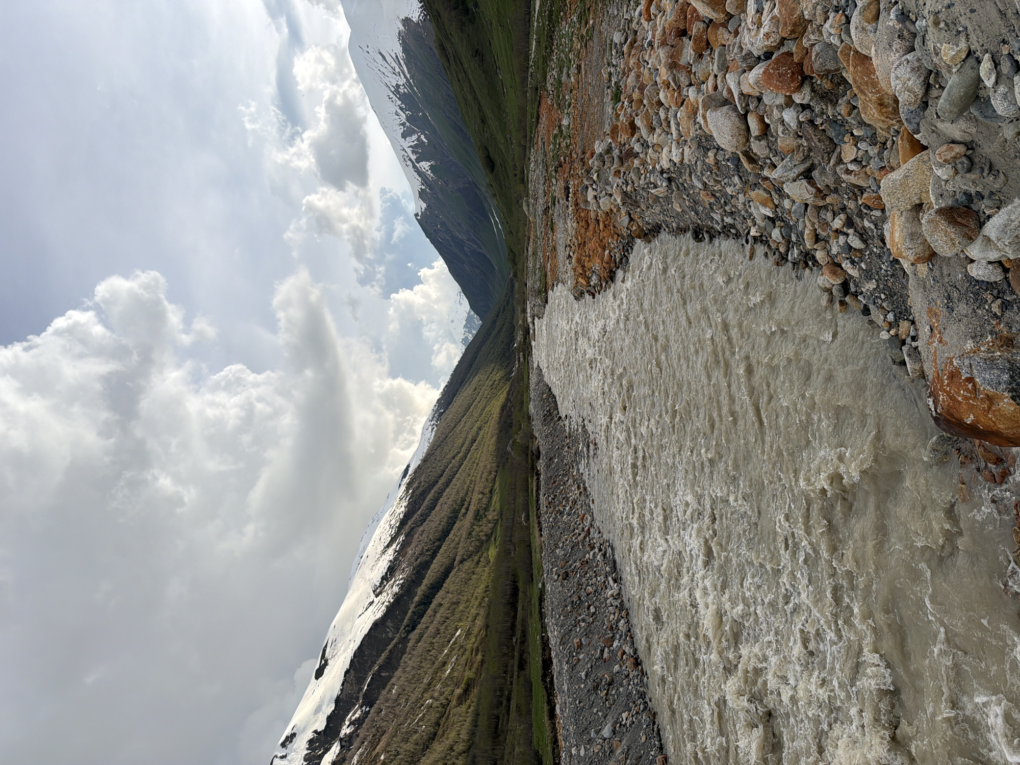 This image depicts a wide, fast-flowing river in a mountain valley, likely located in Ushguli, Georgia. The river's water is murky and appears to be carrying sediment, with white foam indicating turbulence. The riverbanks are covered with a mixture of grey and reddish-brown rocks of various sizes, with some finer gravel and sand visible.
The valley slopes are covered in a mix of green vegetation and sparse trees, with patches of snow on the upper reaches of the mountains. The mountains themselves are steep and rugged, with prominent snow-capped peaks in the background. The sky is partly cloudy, with large, white clouds casting shadows on the landscape. There are no people or animals visible in the frame. The scene appears to be a natural, undeveloped landscape, suggesting a remote mountain environment.