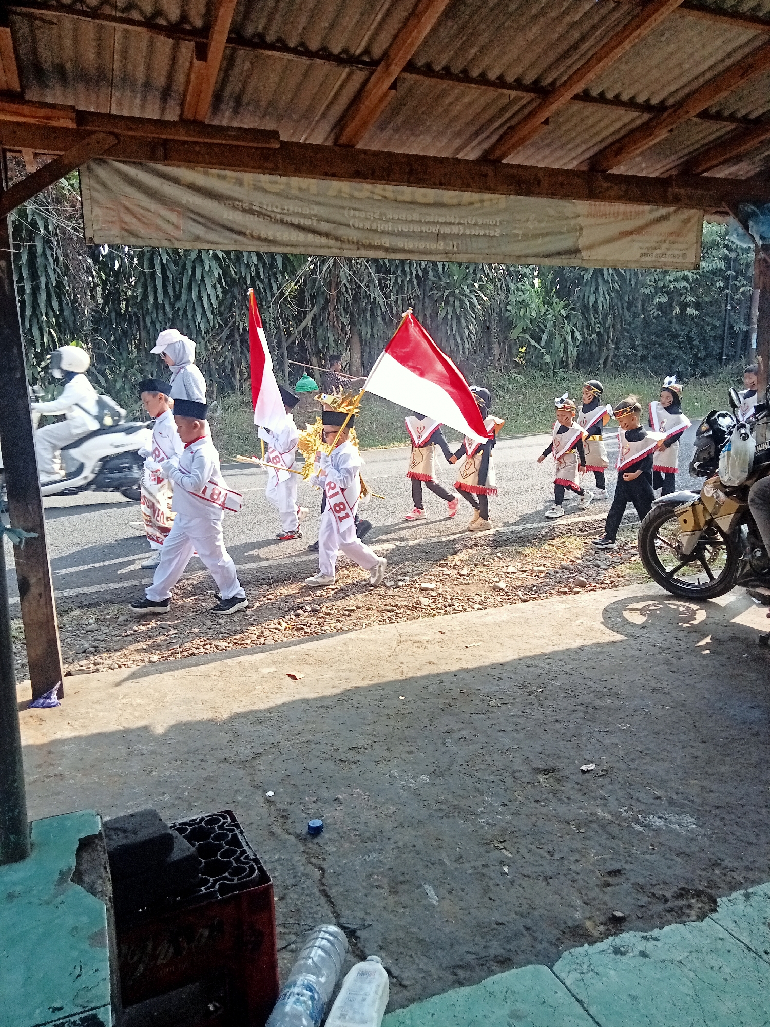 A group of children in uniform, some carrying Indonesian flags, march on a street in Dororejo, Indonesia. The children are wearing white outfits with red and white sashes. The flags are red and white, with one large flag prominently displayed. A motorcycle is parked on the right side of the image. The background consists of trees and vegetation. The scene appears to be outdoors, with a weathered corrugated metal roof structure partially visible in the upper portion of the image. The ground is a mix of dirt and asphalt.