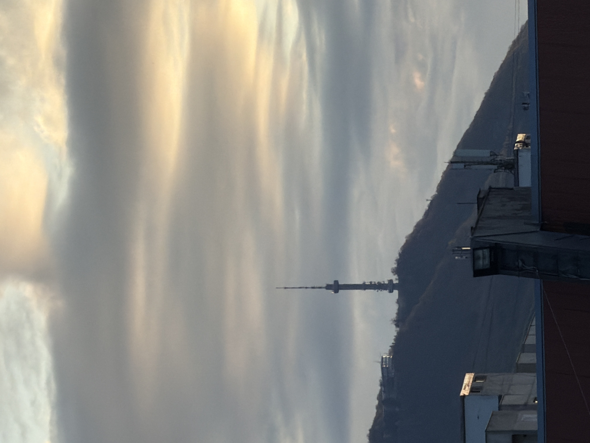 The image shows a view of the Kopitoto TV tower in Sofia, Bulgaria, from a high vantage point. The tower stands silhouetted against a cloudy sky, with a mountain visible behind it. A building is in the foreground, partially obscuring the view.