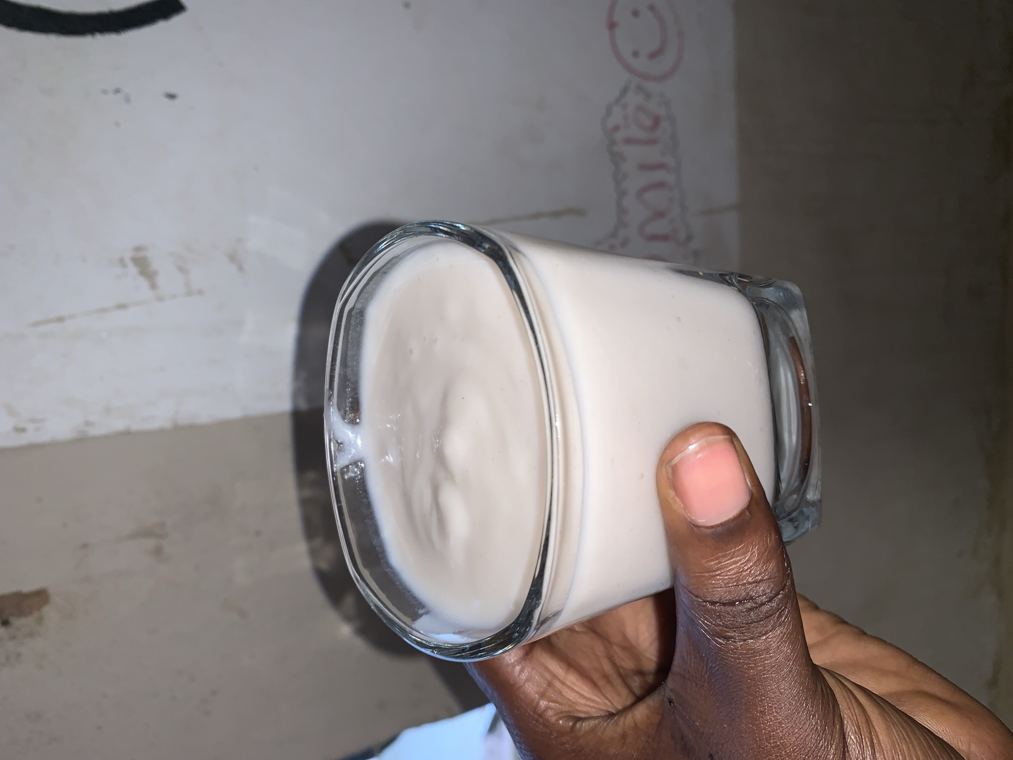 A dark-skinned hand holds a clear, square-shaped glass filled with a white, creamy liquid, likely a beverage, against a backdrop of walls. The walls appear to be in a state of disrepair or unfinished construction, with visible imperfections and what may be discoloration or staining. A red inscription, possibly a child's drawing or writing, is faintly visible on one wall. The location is Rafin Par, Nigeria. There are no other people, subjects, objects, actions, or interactions immediately evident. The environment is dimly lit, adding to the visual texture of the walls.
