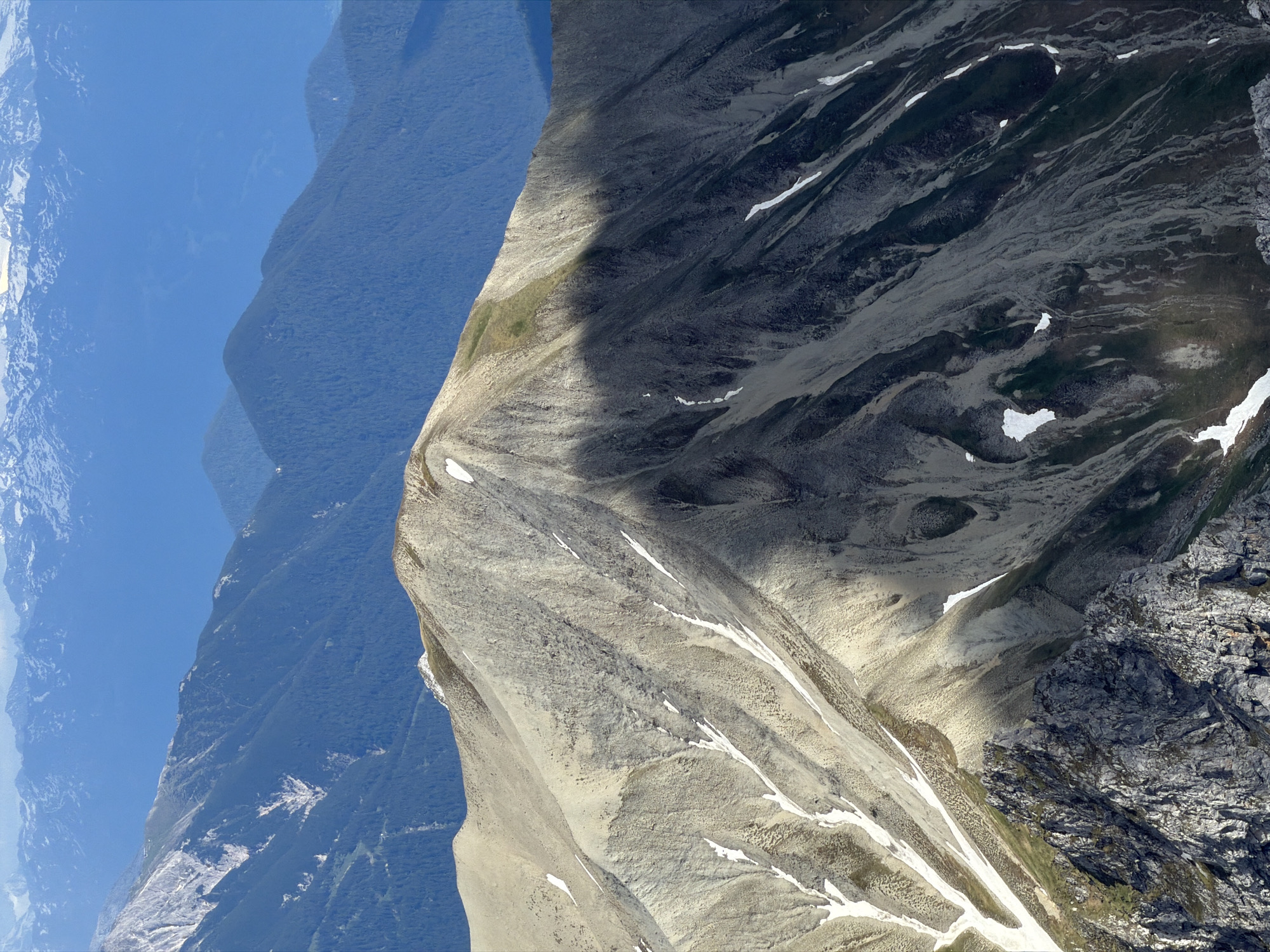 This is a high-angle, telephoto shot of a mountainous landscape with multiple layers of peaks under a clear blue sky. In the foreground, a jagged, rocky outcropping dominates the lower left corner, with patches of green vegetation. Immediately behind and above this, a large, pale-colored mountain slope rises, marked by streaks of snow and shadows. This slope is characterized by a light, gravelly texture. Further back, darker, forested mountains recede into the distance, their slopes appearing steeper and covered in dense trees. The furthest mountains are lighter in color and appear snow-capped, suggesting they are at a higher altitude or further away. The image appears to be a natural landscape, likely in a mountainous region, without any visible structures or people. The city of Garmisch-Partenkirchen, Germany, is known for its proximity to the Alps, making it a plausible location for such a mountainous vista.
