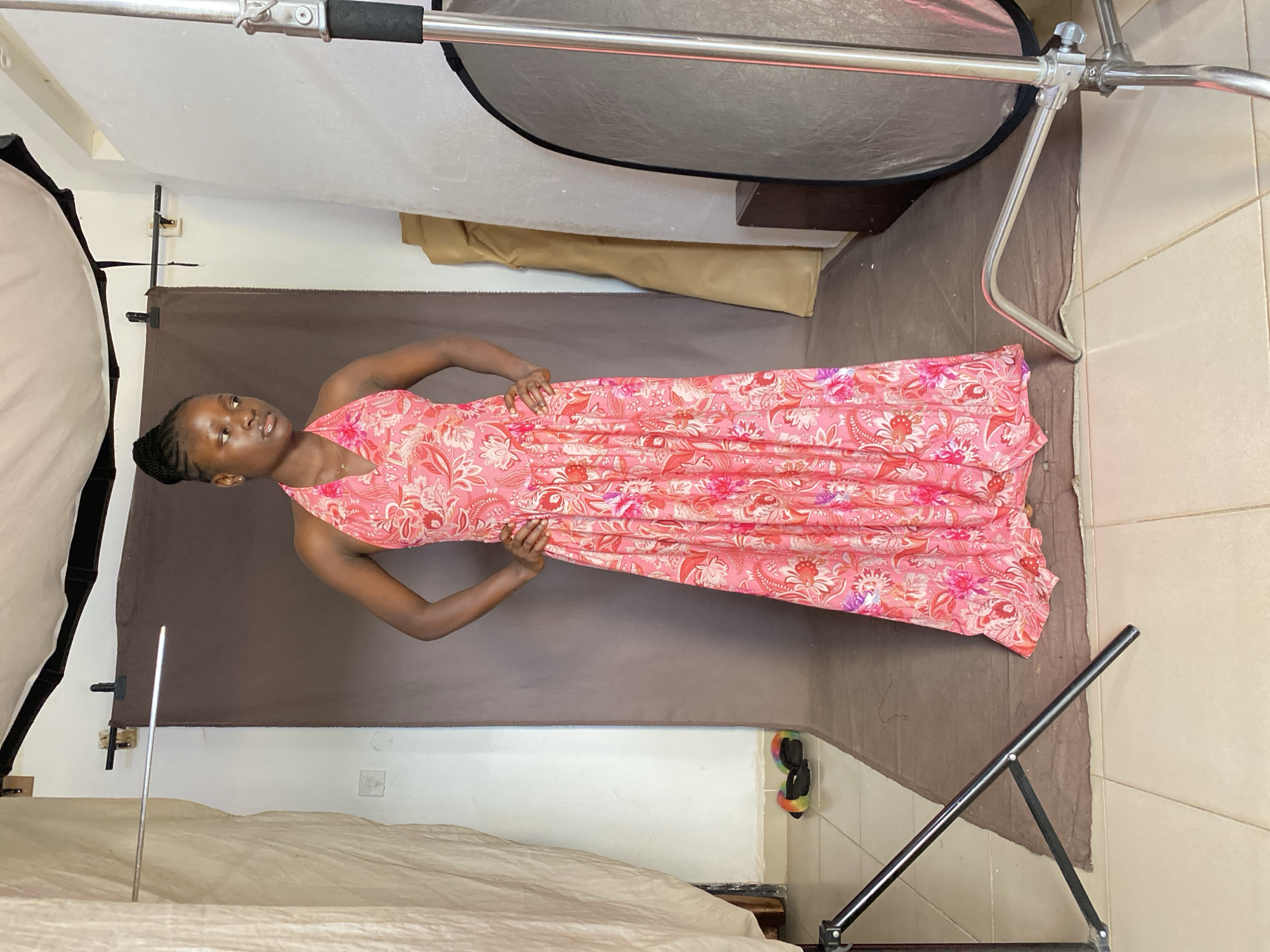 In a room in Bukuru, Nigeria, a young woman stands centered in front of a brown backdrop. She wears a long, pink, floral-patterned halter dress with her hair styled in a bun. Her hands are placed on her hips as she looks to her right. The room appears to be a makeshift photo studio, with photography equipment including a light stand, a reflector, and draped fabric visible. Colorful slippers are on the floor to the left.
