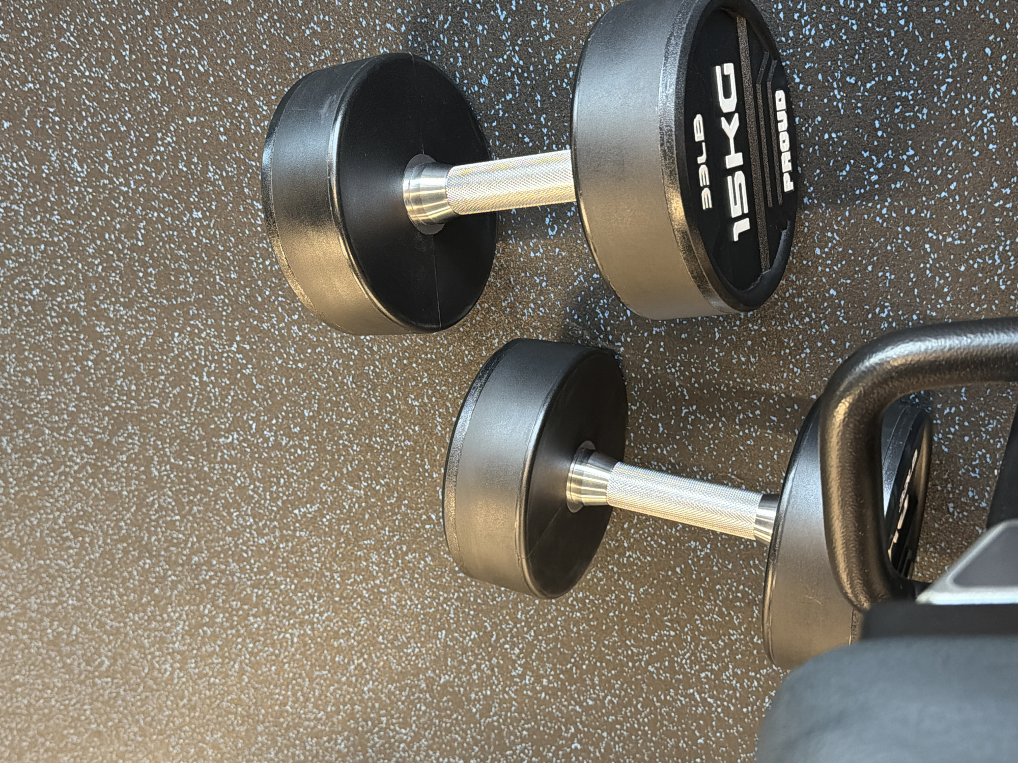 The image shows two dumbbells on a speckled floor. The top dumbbell is black and silver with the weight labeled as 15KG/33LB. The word "PROUD" is also visible on the weight. The second dumbbell is partially visible in the bottom right corner. The setting is a gym floor. The location is unknown.