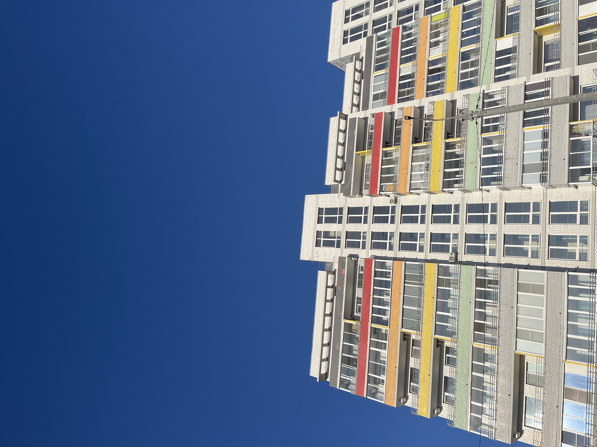 A low-angle shot shows a modern apartment building in Chaikovsky, Russia, set against a clear, deep blue sky. The building is primarily white with evenly spaced, rectangular windows. Vertical columns of red, orange, yellow, and green accent the building's façade. The structure appears to be tiered or stepped, with each level slightly receding. There are no people visible, and the focus is solely on the architecture and its contrast with the sky.