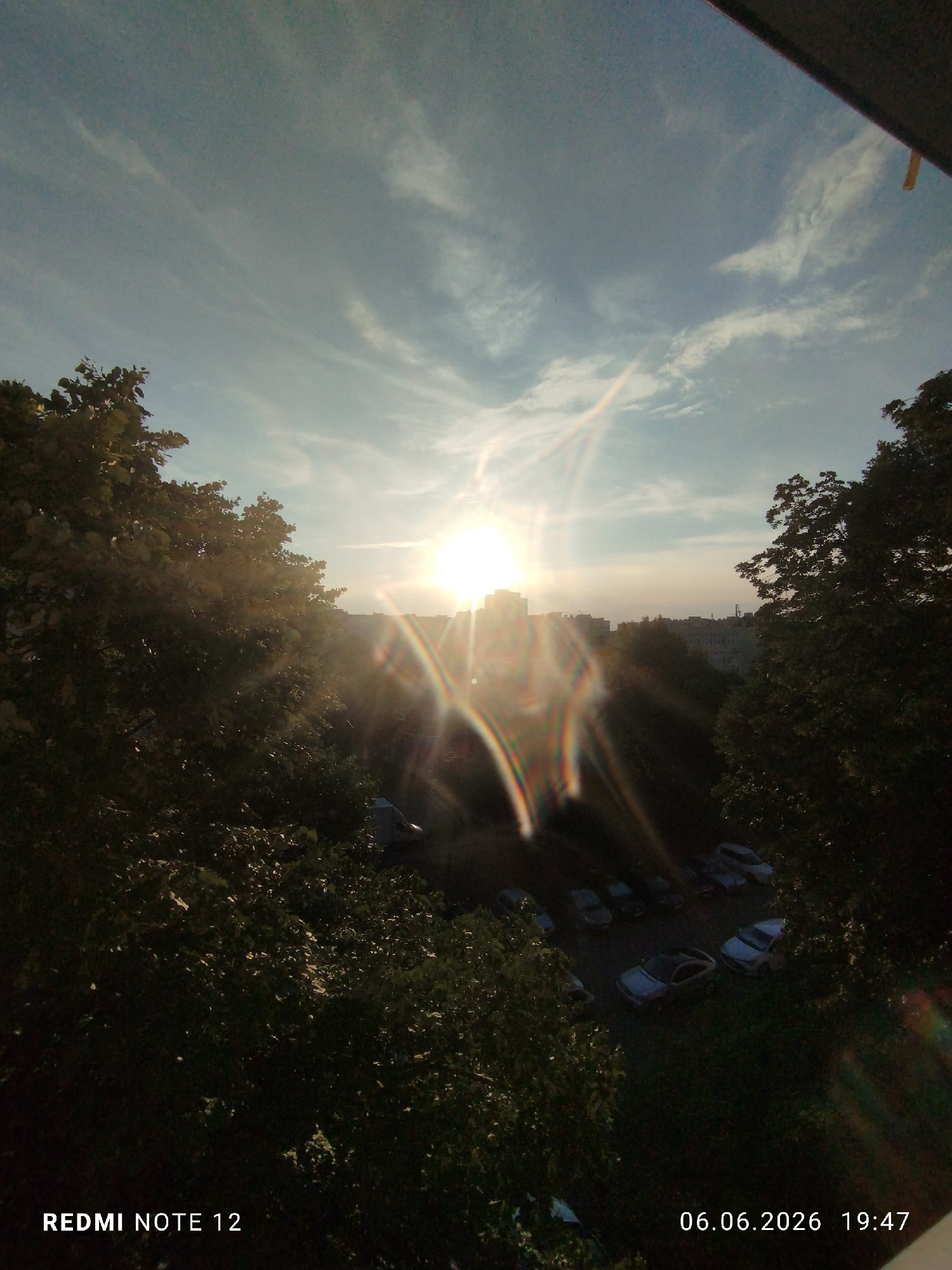 The image captures an outdoor urban scene during the late afternoon or early evening, specifically in the city of Burgas, Bulgaria. The perspective is elevated, looking down through a foreground of dense tree foliage towards a distant urban landscape and a parking area.
The upper portion of the frame features a light blue sky with scattered, wispy white clouds. The sun is a dominant light source, positioned in the upper-mid right, partially obscured by the distant horizon line of buildings. Its intense brightness generates a significant sunburst effect and prominent lens flares, visible as multiple concentric, spectral arcs of color radiating from the light source.
The foreground is heavily framed by the dark, dense canopy of large trees, primarily on the left and right sides. The foliage is severely backlit, appearing in deep shadow with minimal discernible detail.
In the mid-ground, below the sun and between the trees, a cluster of multi-story urban buildings forms a silhouetted skyline against the brighter horizon. Architectural specifics are obscured by the backlighting.
At ground level, partially visible through the trees, is a parking area or street. Several passenger vehicles of various models and colors are parked there, largely obscured by shadows and the surrounding vegetation. A white delivery truck with a blue cargo box is also identifiable on the right side of this ground-level section.
No individuals are discernible within the frame. No specific actions or interactions are depicted.
Embedded technical data in the lower left corner indicates the capture device as "REDMI NOTE 12". In the lower right corner, the timestamp "06.06.2026 19:47" is present.