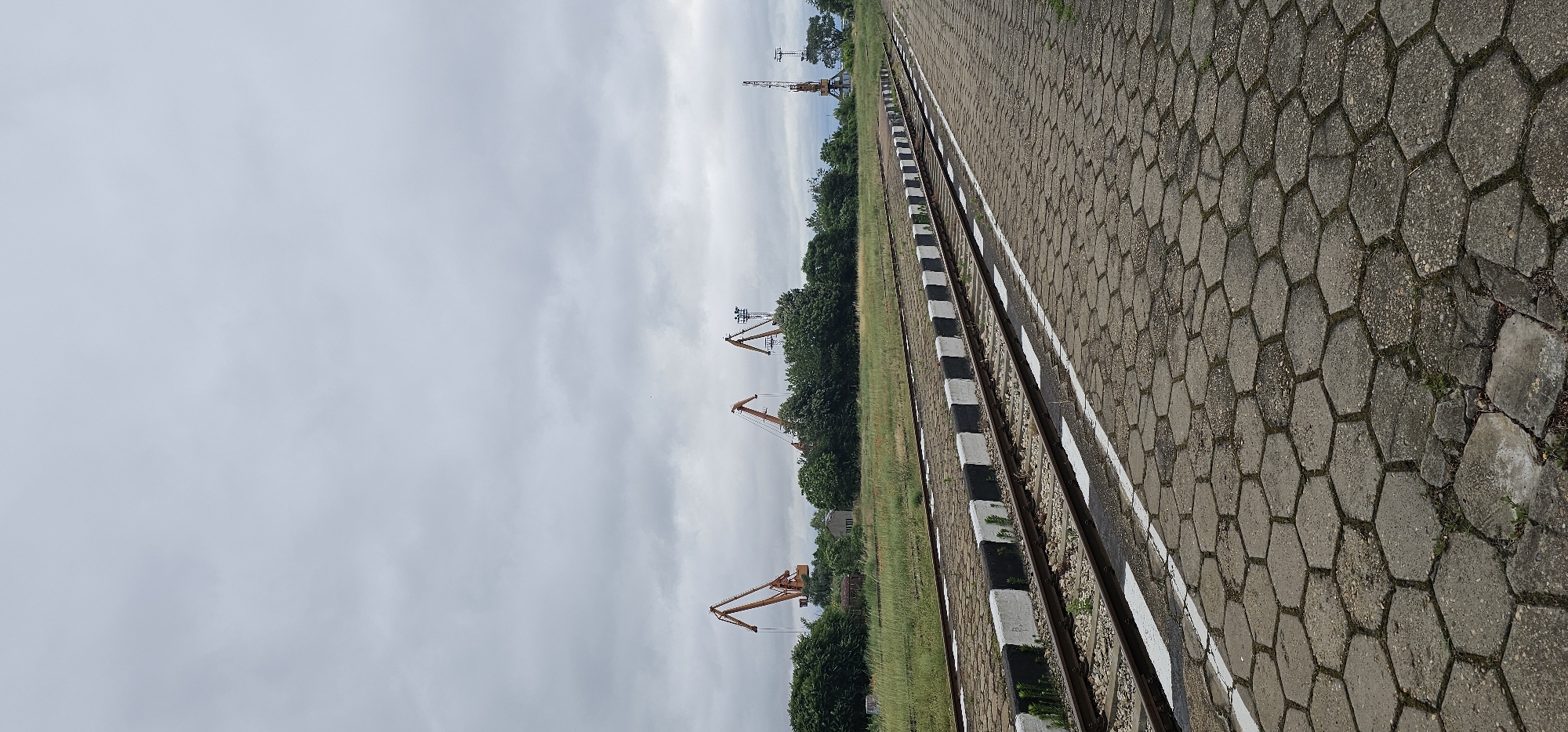 The image shows a view of train tracks and a paved area in the city of Svishtov, Bulgaria. In the background, several large orange cranes are visible above a line of trees. The sky is overcast with gray clouds. The train tracks are on the left side of the image, with a paved area on the right. The paving stones are hexagonal in shape. The scene appears to be an industrial or port area.