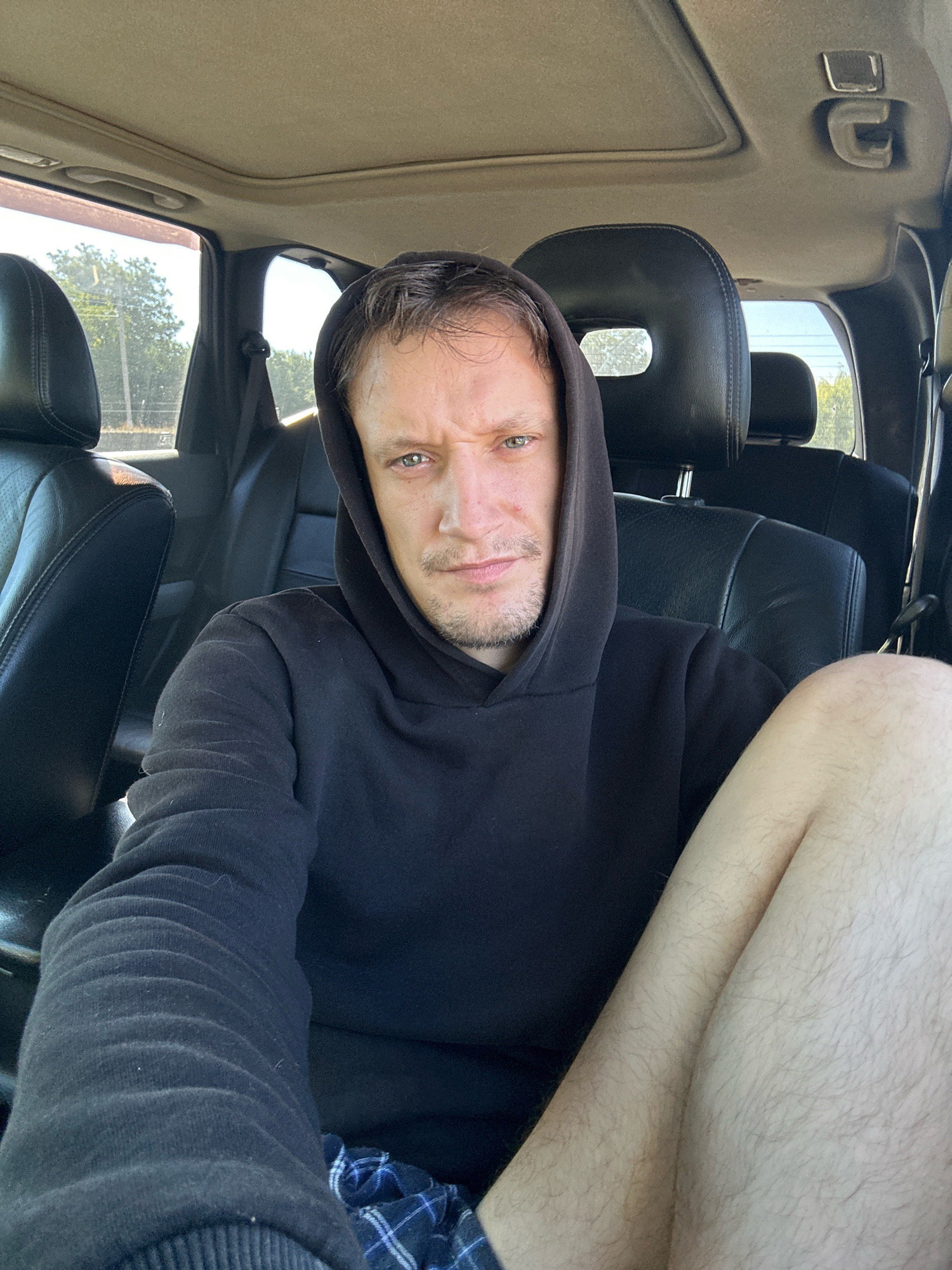 The image shows a man sitting in the driver's seat of a vehicle, likely a subcompact SUV. He is wearing a black hoodie and plaid shorts, and is looking directly at the camera. His left leg is bent at the knee and his right leg is extended. The interior of the car is visible, including black leather seats, the car's headliner, and a sunroof. The exterior of the car is partially visible through the windows, showing trees and a bright sky, suggesting it is daytime. There are no explicit indicators of the location within Makiv, Ukraine, beyond the assumption that the vehicle is situated there.