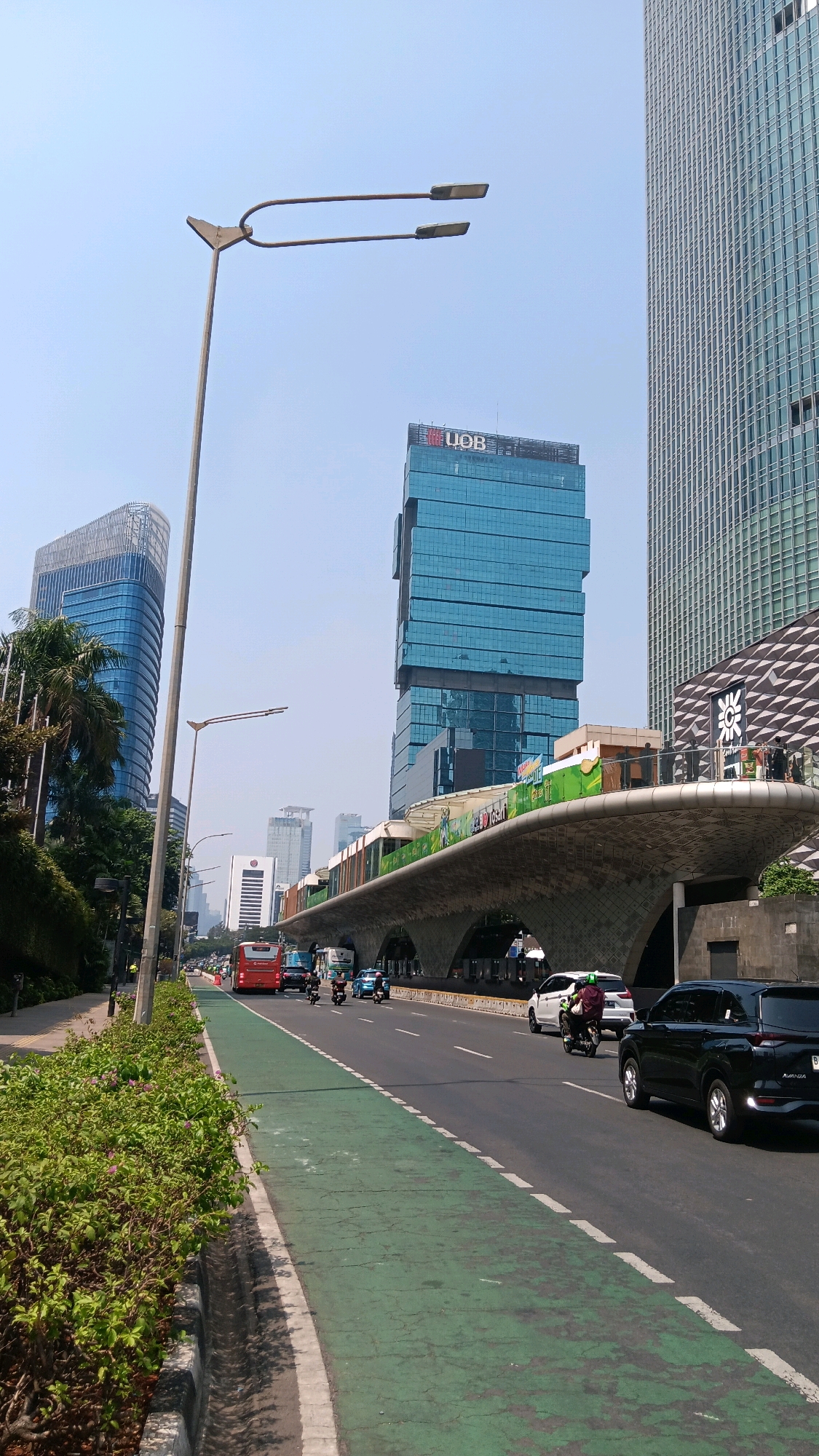 This is a vertical, eye-level, daytime, outdoor, medium shot of a street scene in Central Jakarta, Indonesia. The street is wide and paved with asphalt, with a green lane for bicycles on the left. A row of lush green bushes lines the left side of the street, bordering a sidewalk. In the foreground, a concrete ditch runs along the edge of the sidewalk.
The street is populated by several vehicles, including a red bus in the middle lane, several cars, and a few motorcycles. The vehicles are moving in the same direction, away from the viewer. The sky is clear and blue.
In the background, several modern buildings rise into the sky. The closest building on the right is a tall, glass-fronted skyscraper with a grid-like facade. To its left is another tall building, this one with a distinctive curved architectural design and a lighter-colored facade. Further back, a lighter blue glass skyscraper with a tiered design is visible, along with several other smaller buildings.
An elevated structure, possibly a train or monorail station, arches over the street in the mid-ground. It has a curved, concrete underside with arched openings. The top of the structure is lined with what appear to be advertisements or decorative panels, some featuring green imagery. A few people are visible on a balcony or viewing area of this elevated structure, standing near a patterned facade.
A tall, grey lamppost with two lights is prominently positioned on the left side of the street, in the mid-ground. The scene conveys a sense of urban activity and modern infrastructure.