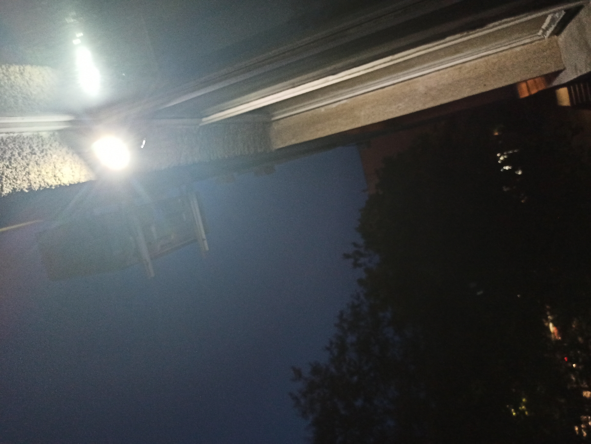 Here is a detailed description of the image:
The image appears to be a nighttime shot taken looking upwards. A building overhang, possibly the roofline, is visible at the top of the frame. The building is constructed of what appears to be concrete and is possibly decorated with white trim. To the right of the building is heavy foliage indicating the presence of trees. A light source, possibly a street lamp, is illuminating the frame with a significant glare to the left of the image and lighting up another building. Beyond the buildings and the light are glimpses of a dark blue night sky.