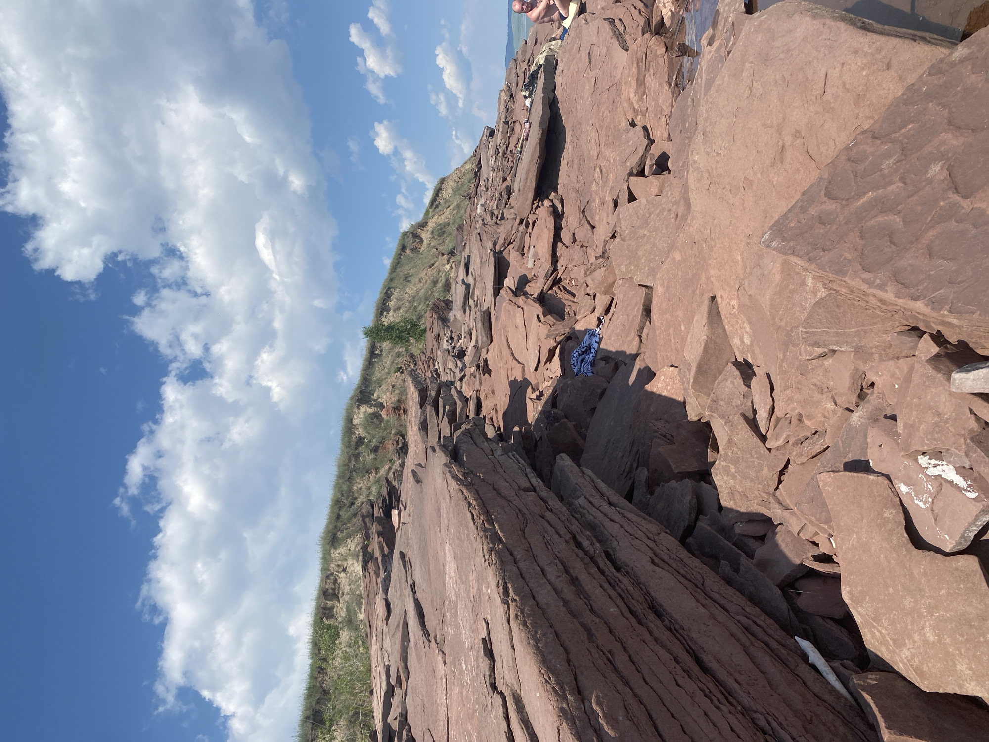 The image depicts a rocky shoreline with a steep, grassy embankment in the background under a bright blue sky with scattered white clouds. The foreground is dominated by large, flat, reddish-brown rocks that appear to be layered. Some smaller rocks and debris are scattered among them. In the mid-ground, a person is seated on the rocks, facing away from the camera, wearing dark shorts and a light-colored shirt. Next to them are some personal belongings, including a bag and a towel. Another towel, blue and white, is draped over a rock closer to the foreground. The scene appears to be a natural outdoor environment, likely a lake or sea shore.