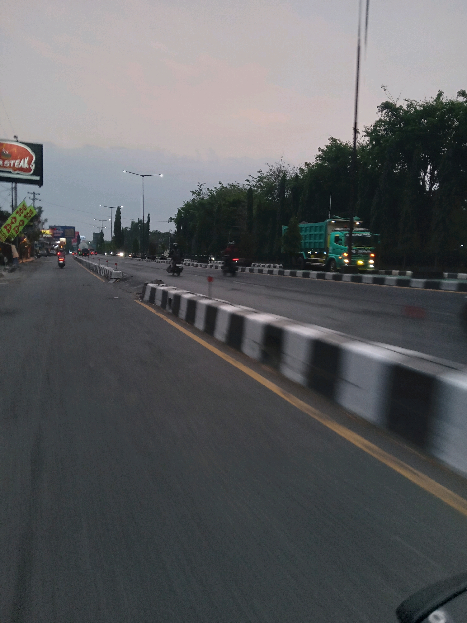 The image depicts a street scene in Kebondalem Kidul, Indonesia. The viewpoint is low, suggesting it was taken from a moving vehicle. The road is paved with asphalt and has yellow lane markings. A barrier with black and white stripes separates lanes on the right side of the road. Trees line the right side of the road, and the sky is overcast with a pale, diffused light.
In the distance, a green dump truck is visible, its headlights illuminated. Several motorcycles are also on the road, with some appearing to be in motion. On the left side of the road, a sign with the word "STEAK" is prominently displayed. Other signs and buildings are also present in the background, indicating a commercial area. Streetlights are visible along the road. The overall atmosphere is that of a moderately busy street at dusk or dawn.