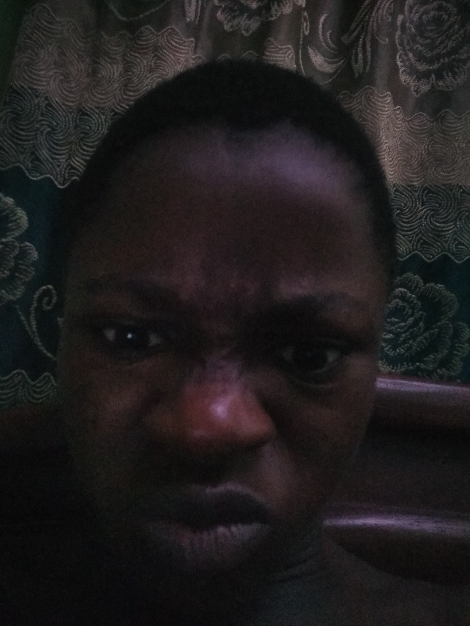 A close-up portrait of a dark-skinned person with a very short haircut, appearing to be in a dimly lit room. The person is looking directly at the camera with a tense facial expression, characterized by a furrowed brow and pursed lips. Behind the subject is a patterned curtain or fabric hanging on the wall. While there is no way to confirm if this picture was taken in Shangisha, Nigeria, based on this picture alone, there are no landmarks or contextual clues in the picture to verify the location.