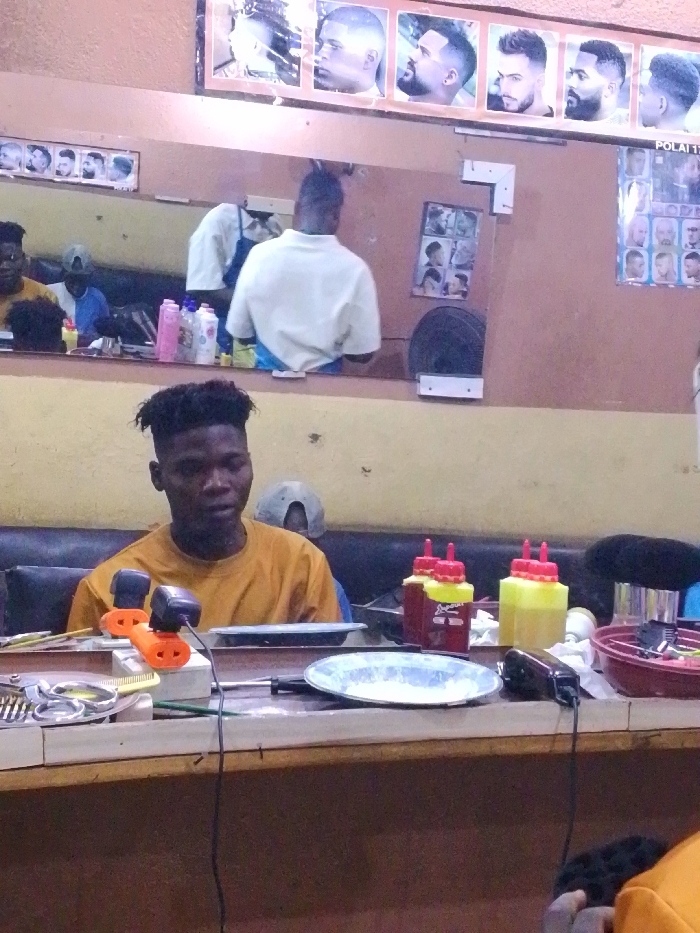 A young man with a high-top haircut wearing a mustard-colored shirt sits in a barber shop. In front of him is a counter with tools, including scissors, clippers, and bottles with red lids. Behind him, a mirror reflects the barber at work. A poster of men's haircuts is displayed above the mirror. The walls are painted a light brown color.