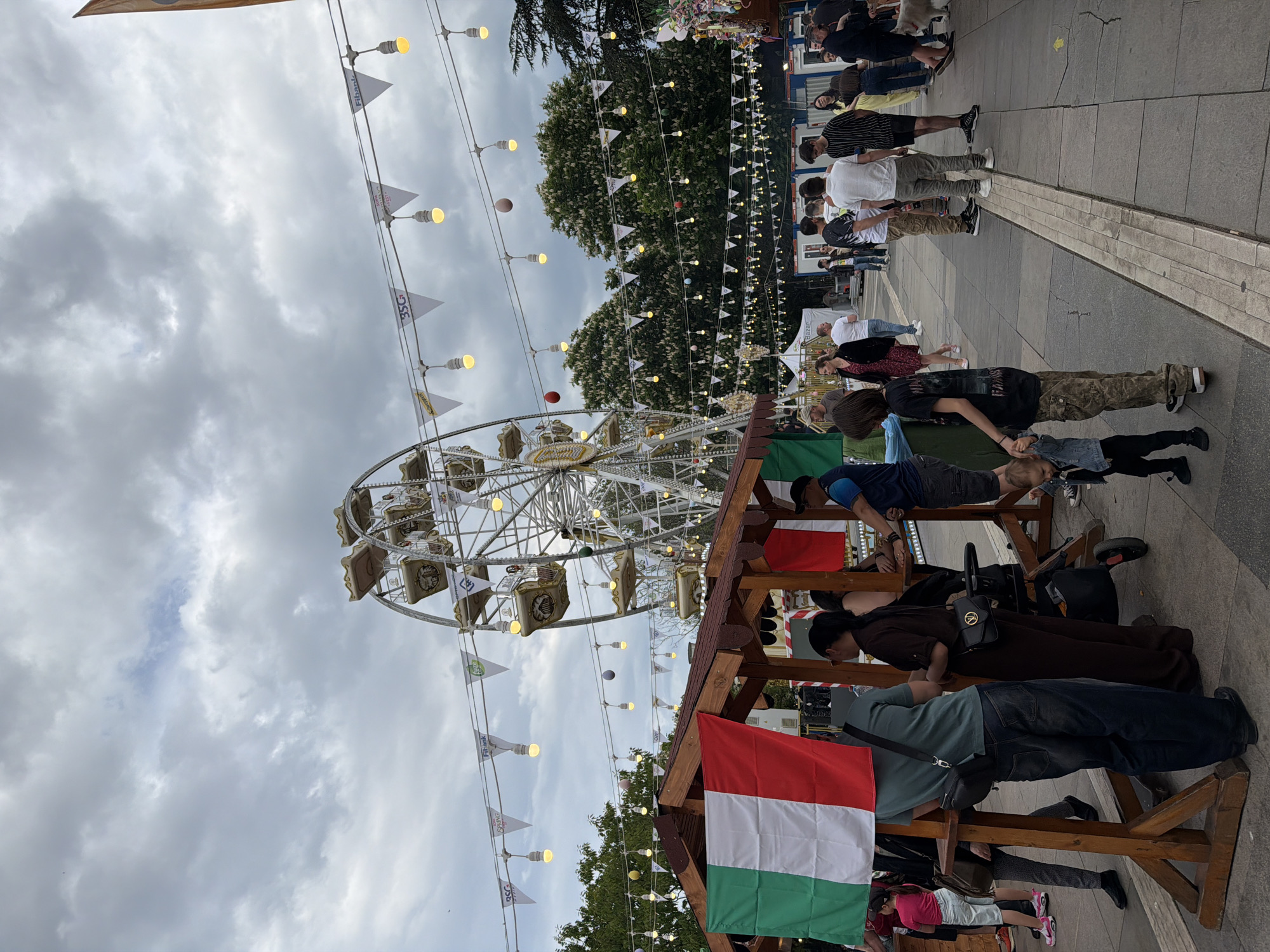 The image depicts a street scene in Sofia, Bulgaria, likely at a fair or festival. A Ferris wheel dominates the background, partially obscured by trees. Strings of lights and pennants hang overhead. In the foreground, a wooden structure is decorated with the flag of Hungary. Several people, including a child, are positioned around this structure. A line of people is visible in the distance, partially obscured by trees. The sky is cloudy. The ground is paved with grey tiles.