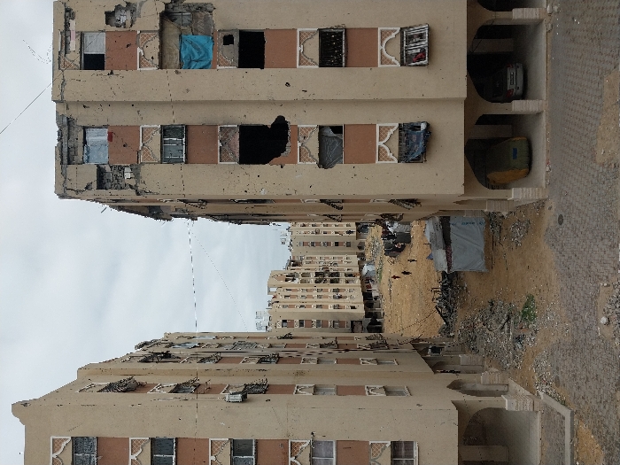 The image depicts a scene in the city of Khan Yunis, Palestinian Territory, showing an urban environment characterized by extensive structural damage. Two multi-story residential buildings, constructed with beige facades and reddish-brown decorative panels, frame a central open area under an overcast sky.
The building on the left exhibits numerous impact holes across its facade and shattered windows, with some window frames missing. The building on the right displays severe damage, including large sections of missing wall revealing interior spaces, widespread impact pockmarks, and broken windows. A blue tarp or fabric hangs from an upper-floor window opening on the right building, and a grey tarp from a lower window. The ground floor of both buildings features arched openings; the right building's arches shelter a white sedan and a large object covered with a yellow tarp.
The central area between the buildings is an unpaved expanse of dirt and substantial rubble, debris, and discarded materials. A rudimentary shelter, covered with a grey tarp, is visible towards the right in this space. In the mid-ground, approximately five indistinct human figures are observed walking across the dirt path. Electrical wires are visible strung between the buildings and across the central open ground. Additional damaged multi-story buildings extend into the background, maintaining a consistent architectural style.