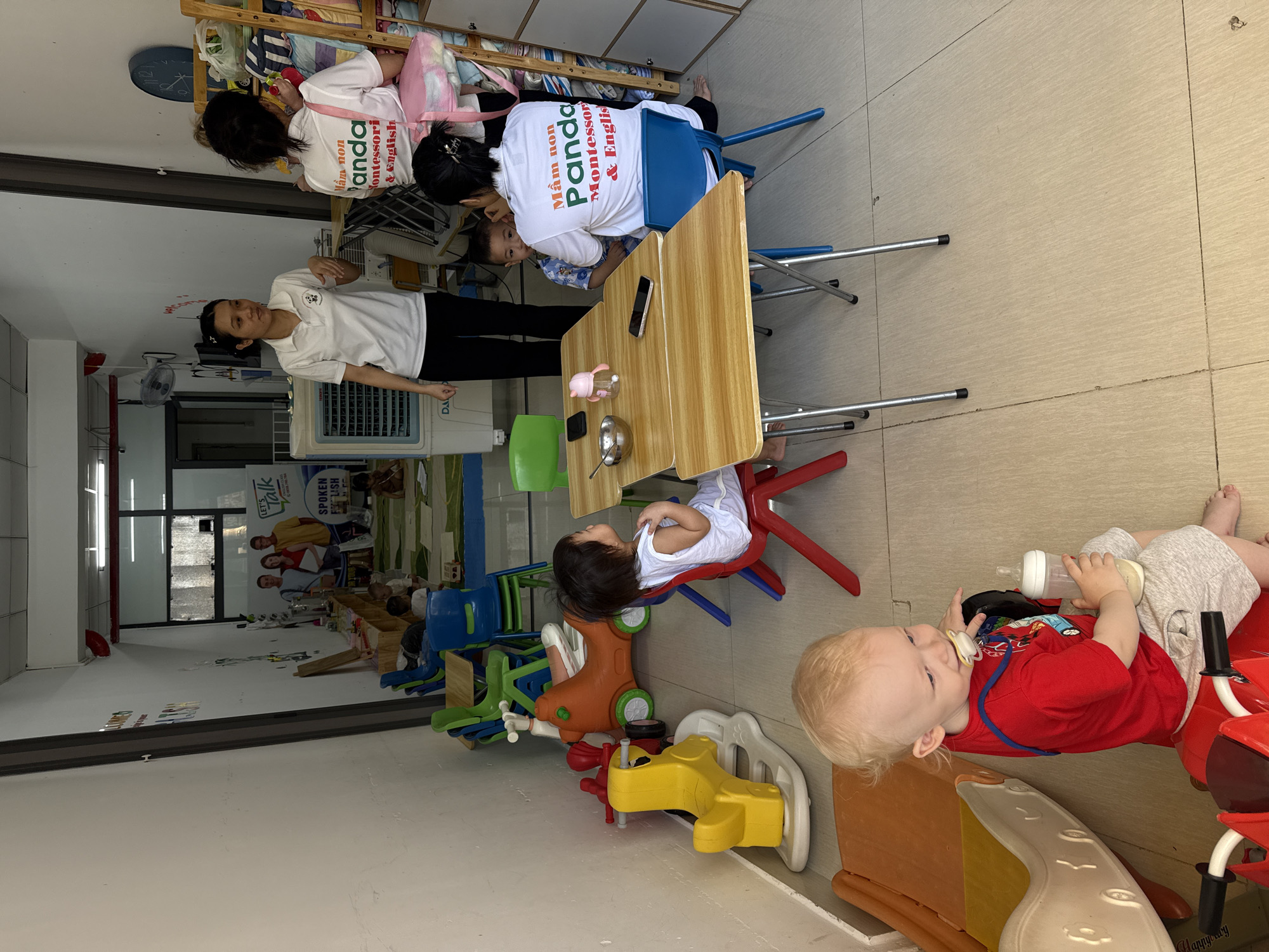 In a brightly lit room with beige tiled floors, likely a daycare or preschool in Nha Trang, Vietnam, several people and children are engaged in various activities. Two adults, dressed in white polo shirts with the text "Mầm non Panda Montessori & English" visible on the back, are interacting with children. One adult appears to be assisting a young child at a light wood table, where a bottle, a bowl, and a phone are placed. Another adult stands nearby, looking towards the table.
Several children are present in the scene. One child, wearing a white top and grey shorts, is lying on a red toy vehicle, holding a bottle and a pacifier. Another child, in a white top, is seated in a red chair at the table. In the background, there are shelves stocked with various items, and an air conditioning unit. Various colorful children's toys, including a red ride-on toy and a yellow toy, are scattered around the room. A large poster with the text "LET'S Talk SPOKEN ENGLISH" is visible on a wall in the background. The overall atmosphere suggests a typical day in an educational setting for young children.