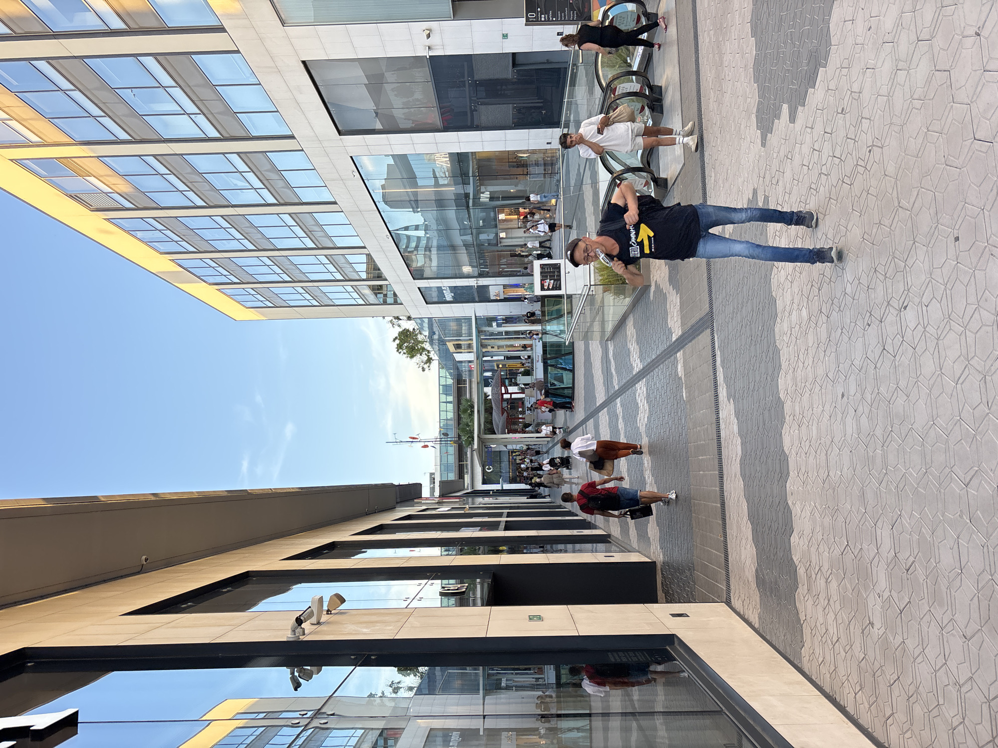 The image displays a lively, sunlit urban street scene in Barcelona, Spain, characterized by modern architecture. The perspective looks down a broad pedestrian avenue bordered by contemporary buildings featuring extensive glass windows that reflect the bright blue sky.
In the foreground, a man wearing a black t-shirt, blue jeans, glasses, and a cap is prominent. He is holding a small bottle, possibly drinking from it. His t-shirt displays a yellow arrow pointing upwards and to the right, with the visible text "CAMINA." Behind him, a man in a white t-shirt and shorts is standing on an escalator, and further up, a woman in black attire is also on an escalator. Numerous other people are visible in the background, walking or entering the various establishments lining the street. On the right side of the street, another man in a red t-shirt and shorts is seen walking away from the camera.
The ground is paved with light-colored, geometrically patterned interlocking tiles, creating a textured surface. The buildings on both sides are tall and sleek, with multi-story glass facades that give a sense of openness and reflect the surroundings. The presence of escalators suggests a multi-level commercial or office complex. The clear, bright sky and distinct shadows indicate a sunny mid-day or afternoon. A faint "ZARA" sign can be seen on one of the storefronts. The scene conveys a sense of everyday urban activity in a modern European city.