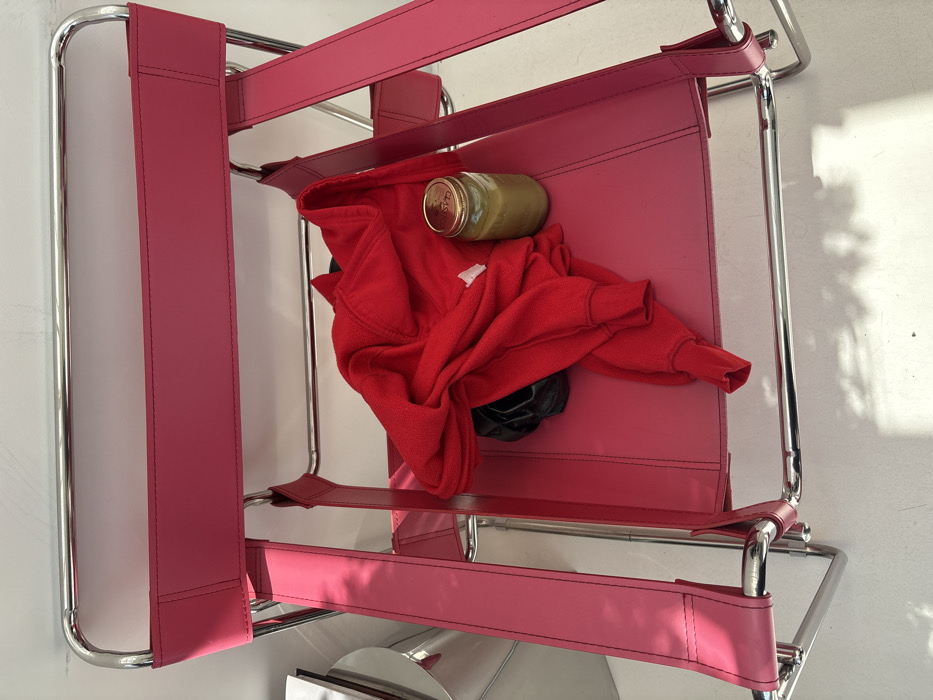 A red and pink chair has a jar, red jacket, and black shoes on the seat. The frame is metal with a hot pink material making up the back and seat. The jacket is bunched up with a portion of the shoes peeking out. The jar is laying horizontally across the top of the chair, partially covered by the jacket. The chair is leaning against a wall.