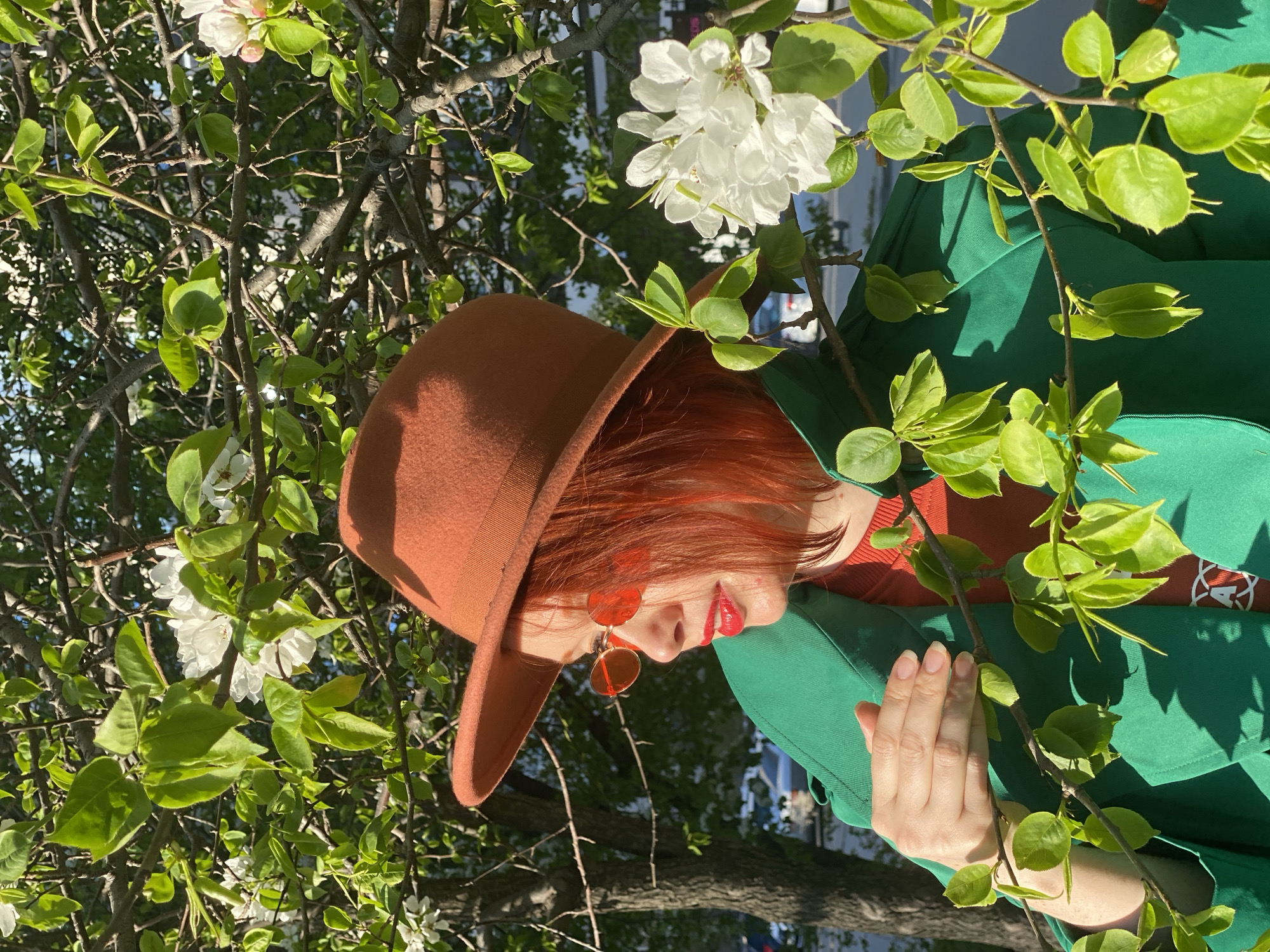 A woman with auburn hair is shown in a medium shot from the waist up. She is wearing a brown fedora, round rose-tinted sunglasses, a green jacket, and a rust-colored shirt. She is standing amidst blooming branches with white flowers and green leaves. Her left hand is raised, with her fingers pointing upwards. The image was taken outside in Yekaterinburg, Russia.