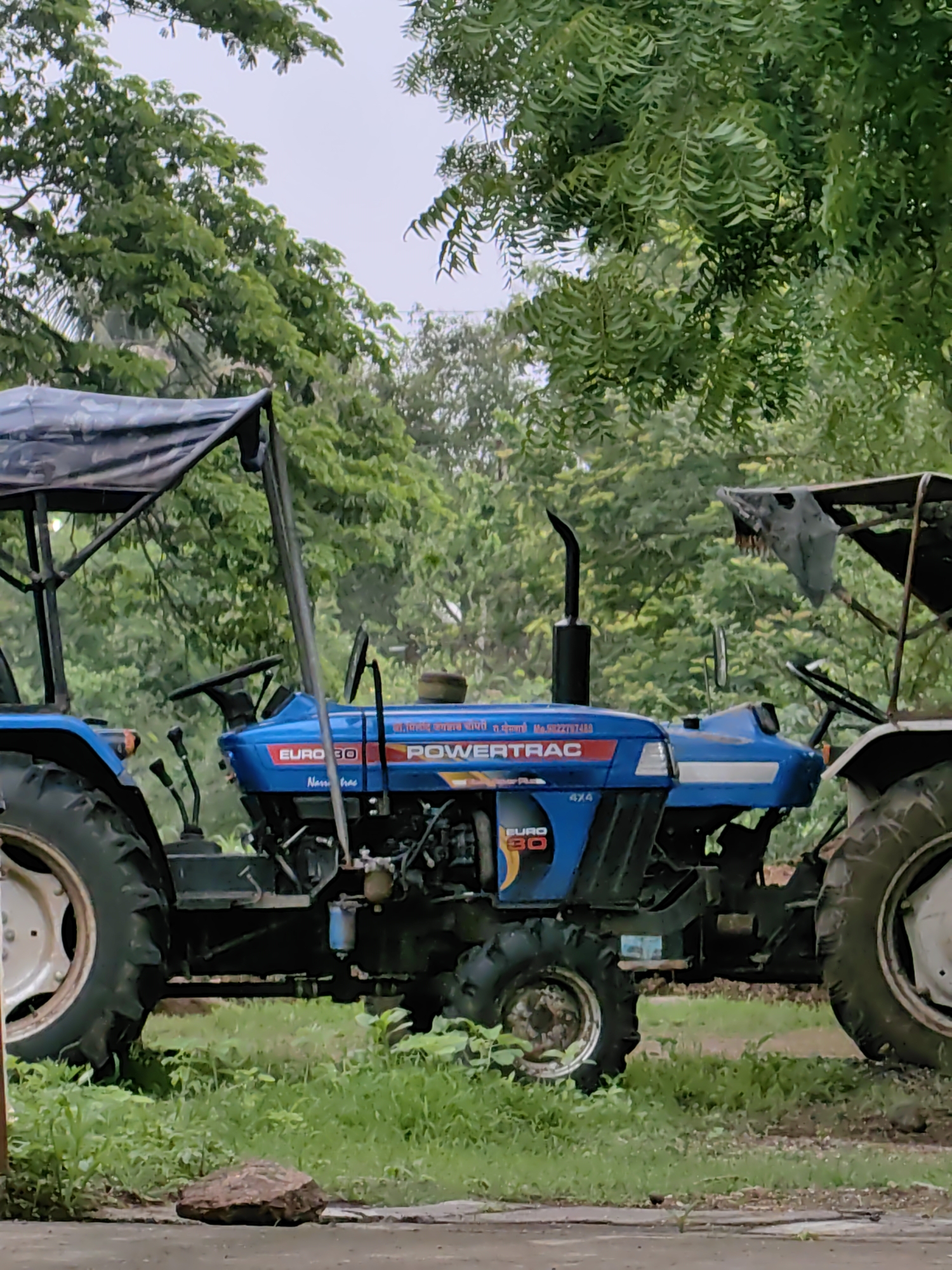 A blue Powertrac Euro 30 4x4 tractor is parked on a dirt and grass surface, possibly in Mhaiswadi, India. The tractor is facing towards the right of the frame. It has a black canopy over the driver's seat, supported by metal poles. The tractor features a large front tire on the left, a steering wheel, and various controls. The body of the tractor has decals with "EURO 30" and "POWERTRAC" in red and white lettering. A smaller rear tire is visible on the left side of the tractor. A portion of another tractor, mostly white with a black canopy, is visible on the far right of the image. The background is filled with lush green trees and foliage, suggesting an outdoor, possibly rural or semi-rural setting. The sky appears overcast.