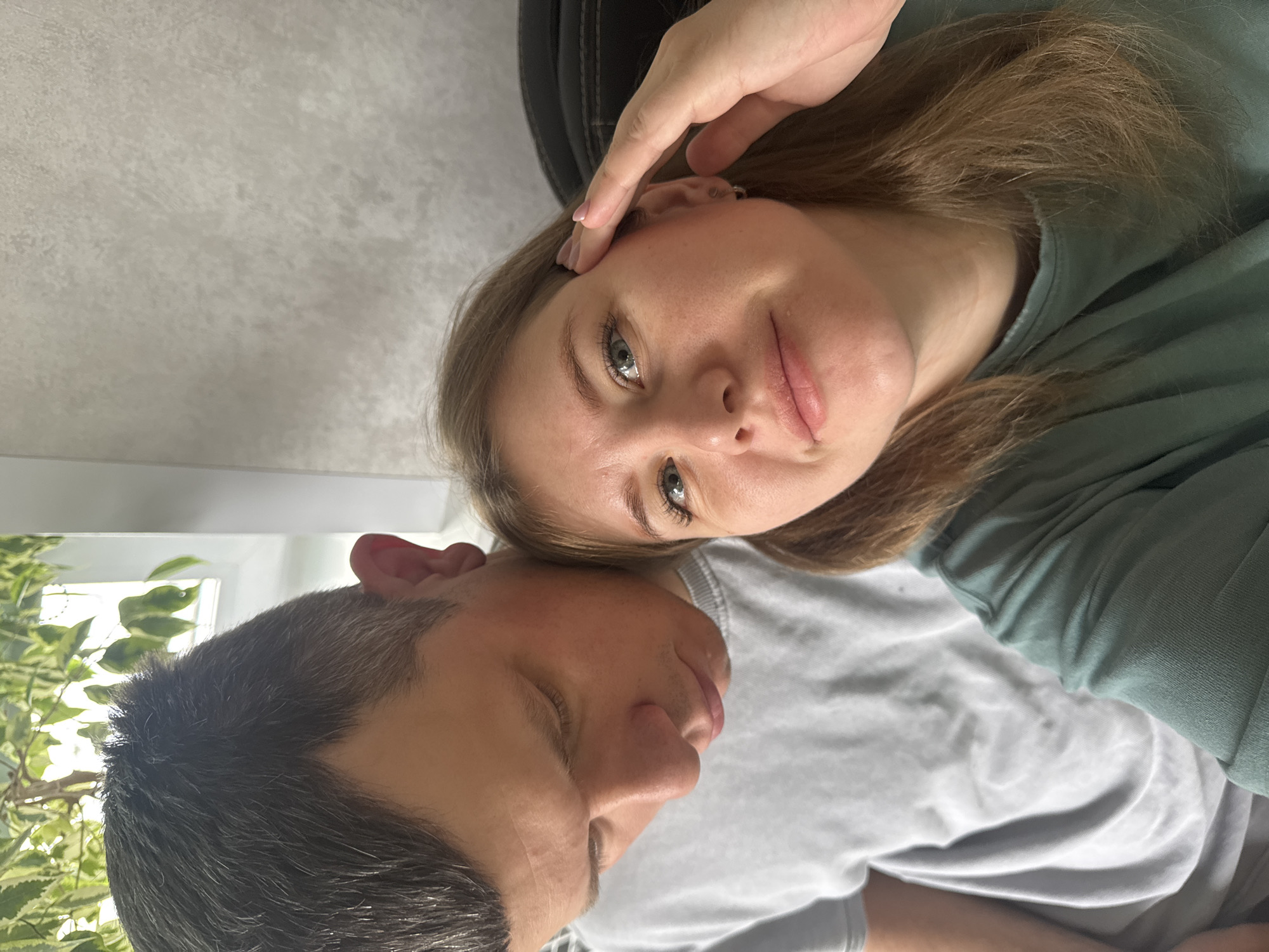 An intimate, close-up indoor image features two individuals, a woman and a man, captured in what appears to be a selfie. The image is oriented with the subjects lying down, or the camera tilted, resulting in a rotated perspective where the ceiling is on the left side of the frame and the floor/lower bodies are on the right.
In the foreground, a young woman with light brown hair and striking blue-green eyes looks directly at the camera with a soft, neutral expression. She wears a teal or dark green top, and her left hand is gently placed near her temple. Adjacent to her and slightly behind, a man with short, dark hair (showing hints of grey) is visible. He wears a light grey t-shirt, and his face is turned away from the camera, resting near the woman's head.
The background elements suggest a domestic setting. The upper left portion of the image reveals a light-colored, subtly textured ceiling or wall. In the lower left, behind the man's head, a lush houseplant with variegated green and white leaves is visible, suggesting proximity to a window and natural light. A dark, possibly upholstered, object is partially seen behind the woman's head.
The close proximity of the individuals conveys a sense of closeness and affection. The lighting indicates it is daytime. The setting is indoors, likely a home in Gornyi (Dvurechenskoe S/P), Russia. There is no visible text in the image.