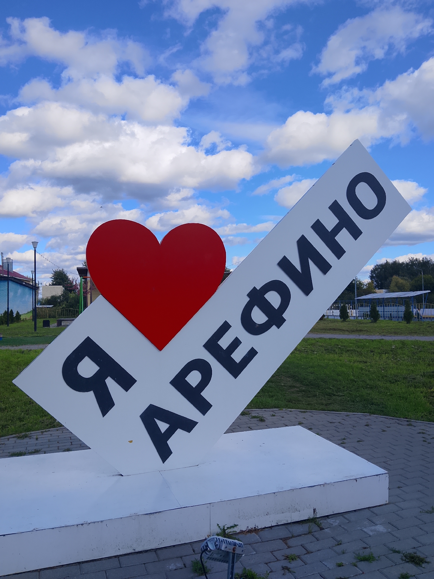 An outdoor monument, featuring a large red heart next to a white sign with dark Russian lettering, is positioned on a paved area. The sign is tilted and reads "Я АРЕФИНО", signifying "I love Arefino." In the background, there are grassy areas, trees, and buildings, suggesting an urban or semi-urban setting. The sky above is blue with scattered white clouds. The monument appears to be a landmark in the city of Arefino, Russia.
