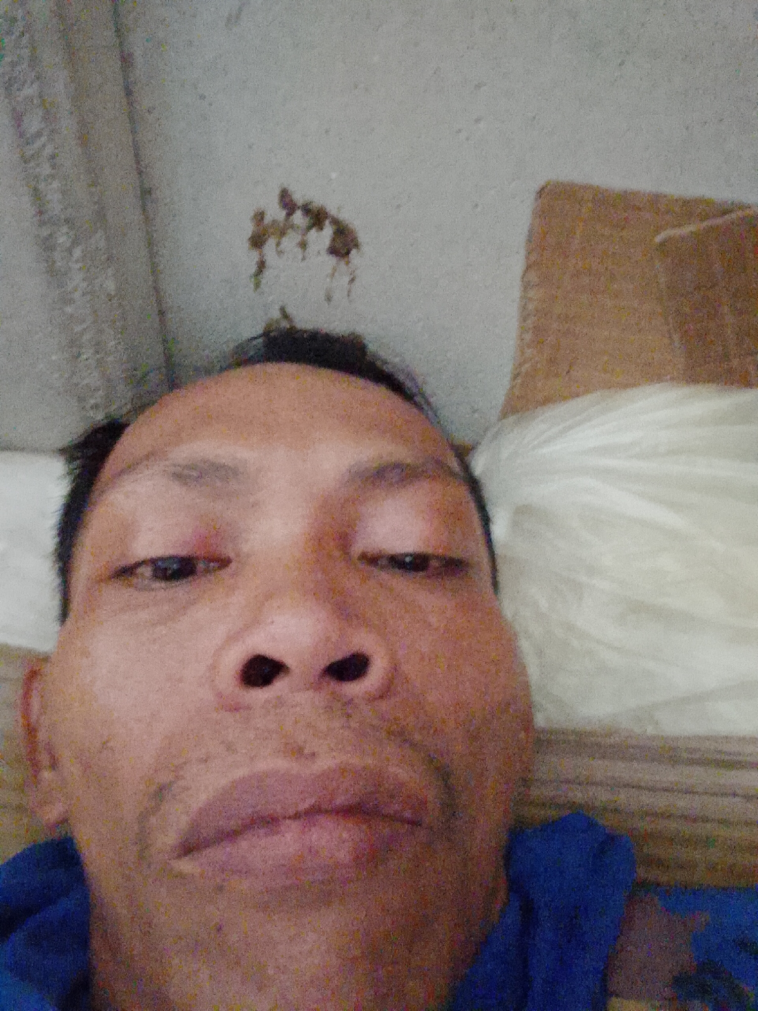 A close-up selfie of a middle-aged Asian man with dark hair and brown eyes. He is lying down on a surface with a white pillow behind his head. He is wearing a blue shirt. The background includes a gray ceiling with a stain near the top of the image. The location is Bukit Ketok, Indonesia.
