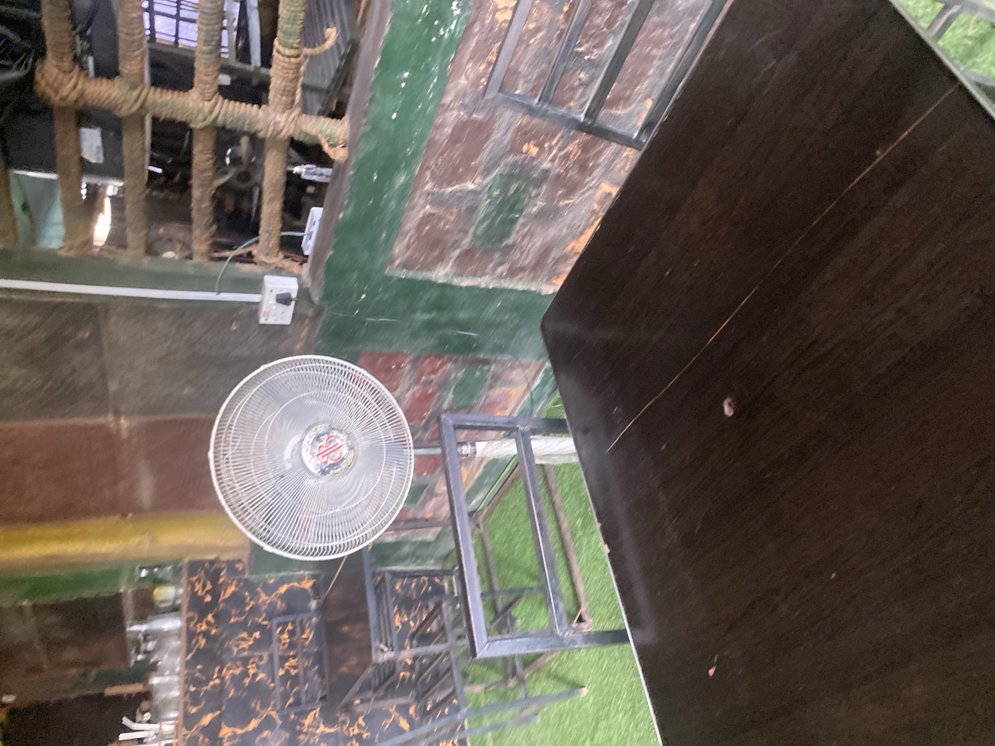 The image depicts an indoor seating area, likely a restaurant or cafe, situated in Jalingo, Nigeria. A dark wooden table dominates the foreground, with its surface showing some minor imperfections. Behind it, a metal-framed chair with a dark seat is partially visible. To the left, another identical chair and table are set up, with a metallic grille and a patterned wall featuring black and orange swirls. In the middle ground, a large oscillating fan stands on a white pedestal, facing towards the tables. The background reveals a textured wall with reddish-brown and green plaster, and a rustic-looking window or partition covered with coarse brown rope. A white electrical socket is mounted on the wall to the left of the fan. The floor beneath the chairs and tables is covered with artificial green turf.
