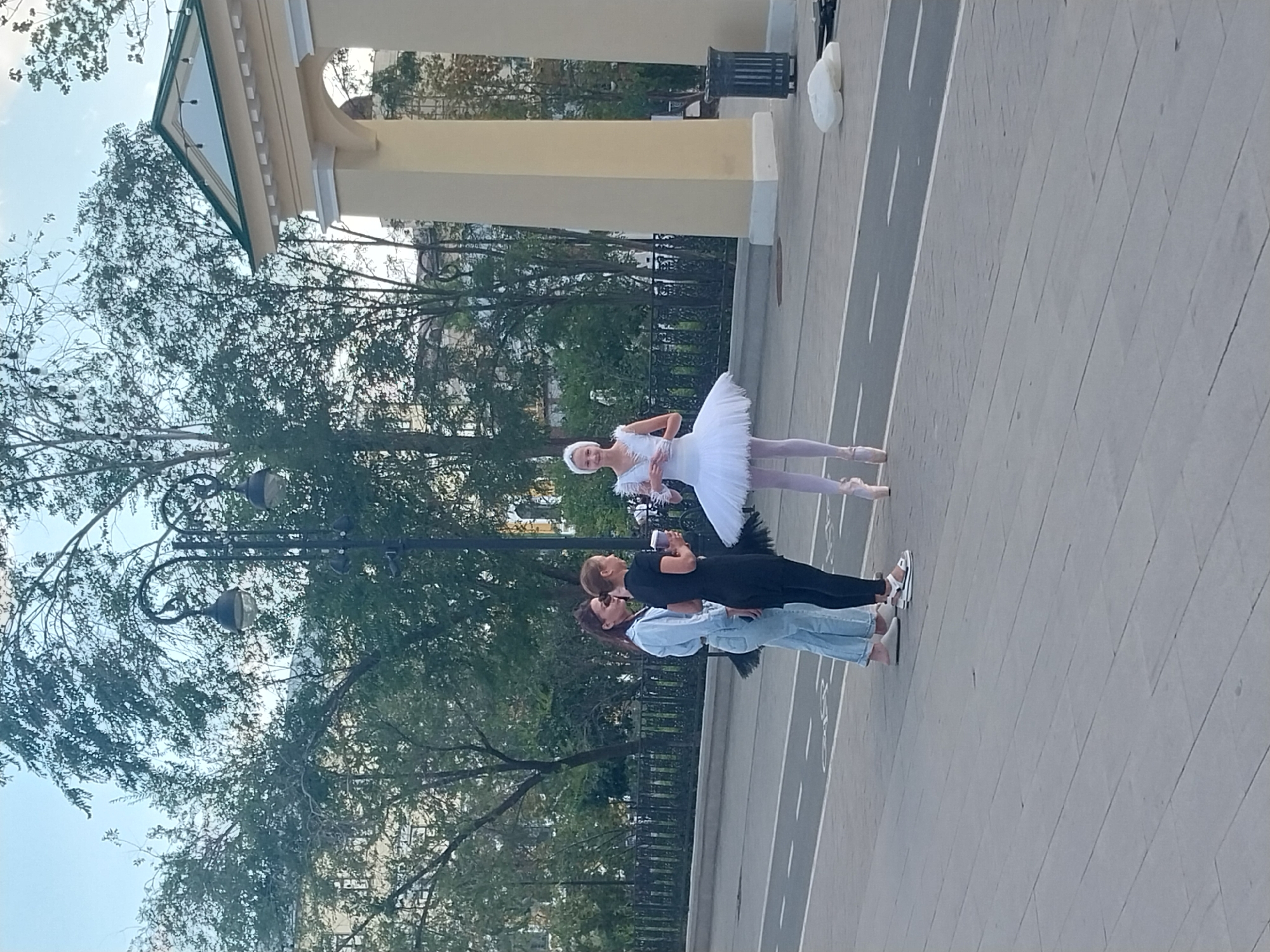 An outdoor daytime scene shows a young woman in a white ballet tutu and pointe shoes standing on a paved area. To her left, two other women stand talking. The woman on the far left is wearing a light blue denim jacket and light blue jeans, while the woman next to her is wearing a black short-sleeved dress and black leggings. The woman in the black dress is holding a paper cup with a lid. Behind the women, a black metal fence runs along a street, with trees and buildings visible beyond the fence. A large, cream-colored architectural structure with columns is to the right of the ballet dancer. A dark metal trash bin is also visible to the right of the structure. The paved area appears to be a pedestrian walkway or plaza, with white lines marking a lane or path on the ground. The scene is set in the city of Orenburg, Russia.