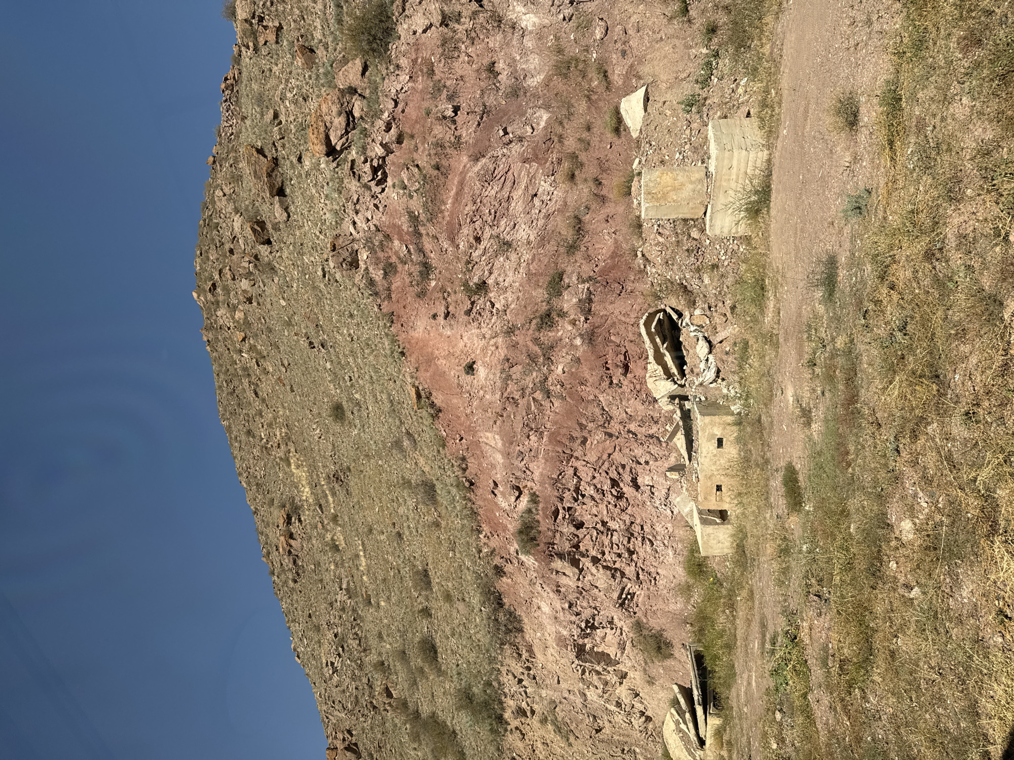 The image depicts a desolate, arid hillside under a clear blue sky. The terrain is characterized by rocky, dry vegetation, and a prominent reddish-brown geological stratum cutting across the slope. In the foreground, there are remnants of concrete structures, appearing as weathered blocks and what looks like a partially constructed or ruined building with two small rectangular openings. Scattered concrete debris is also visible, along with patches of dry grass and sparse, hardy vegetation. The overall impression is one of a remote, perhaps abandoned, or undeveloped area, consistent with a location in the outskirts of Sovetskoye, Kyrgyzstan.