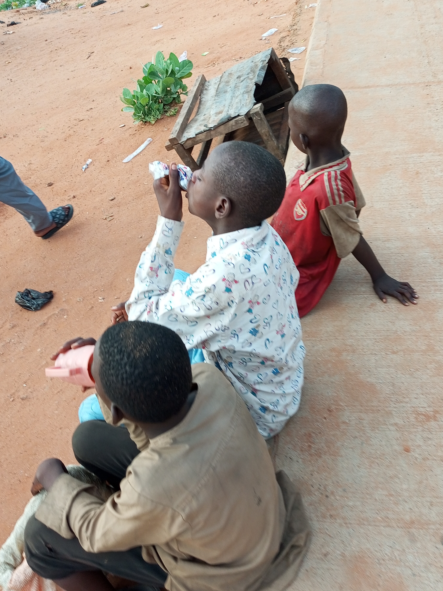 The image depicts three young boys in an outdoor setting, likely in the city of Gombe, Nigeria. The ground is composed of reddish-brown earth, common in the region, with a strip of concrete running along the right side of the frame. In the background, a small, makeshift wooden structure with a rusted metal roof sits on the sandy ground, near some sparse green vegetation.
The boy in the foreground, viewed from behind, wears a light brown long-sleeved shirt and dark pants, and is holding a pink cup. To his left, another boy, facing away from the camera, is dressed in a white shirt with a repeating pattern of small blue and pink figures. He is holding a white object up to his mouth, appearing to drink or eat from it. A third boy, also facing away, is positioned to the right, wearing a red shirt with grey accents and a small crest. His arm is extended, resting on the concrete surface. The legs of another individual wearing grey pants and dark sandals are visible on the far left of the image, suggesting their presence just outside the frame. Scattered on the ground are small pieces of litter, including a crumpled black plastic bag.