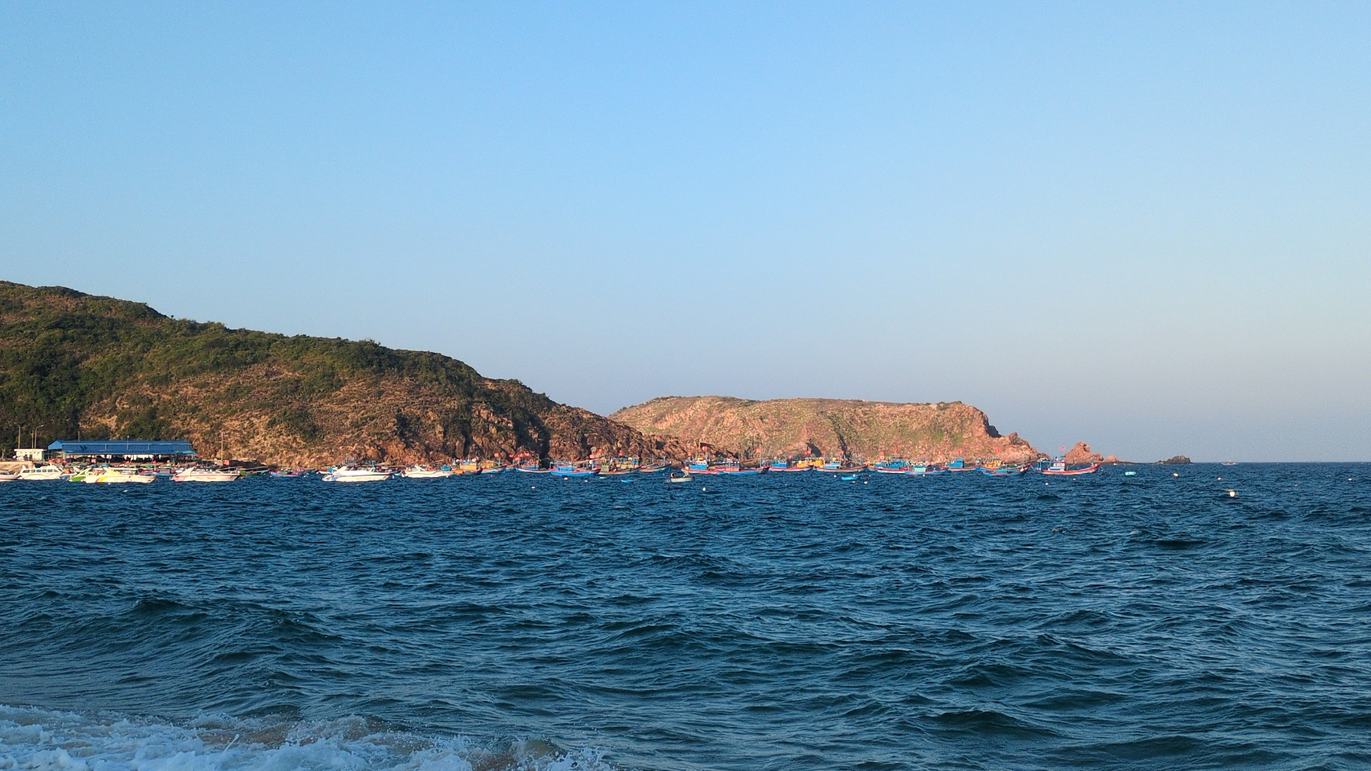 The image displays a coastal scene in East Quy Nhon Ward, Vietnam, characterized by a clear blue sky over a deep blue sea. The foreground shows choppy ocean water with white foam from a breaking wave at the bottom left. In the midground, a dense line of approximately fifty to sixty boats is moored or anchored close to the shoreline. These include numerous traditional Vietnamese fishing boats with blue hulls and accents of red, yellow, and green, interspersed with several white speedboats or smaller recreational vessels. The background features a rugged landscape composed of multiple hills. To the left, a large, steep mountain is densely covered in green vegetation. At its base, a structure with a prominent blue roof is visible near the water. Towards the center and right, rocky, brown hills with sparse vegetation descend towards the sea, with some rock formations extending into the water. The sky is uniformly clear and light blue, suggesting fair weather. No individuals are explicitly visible.