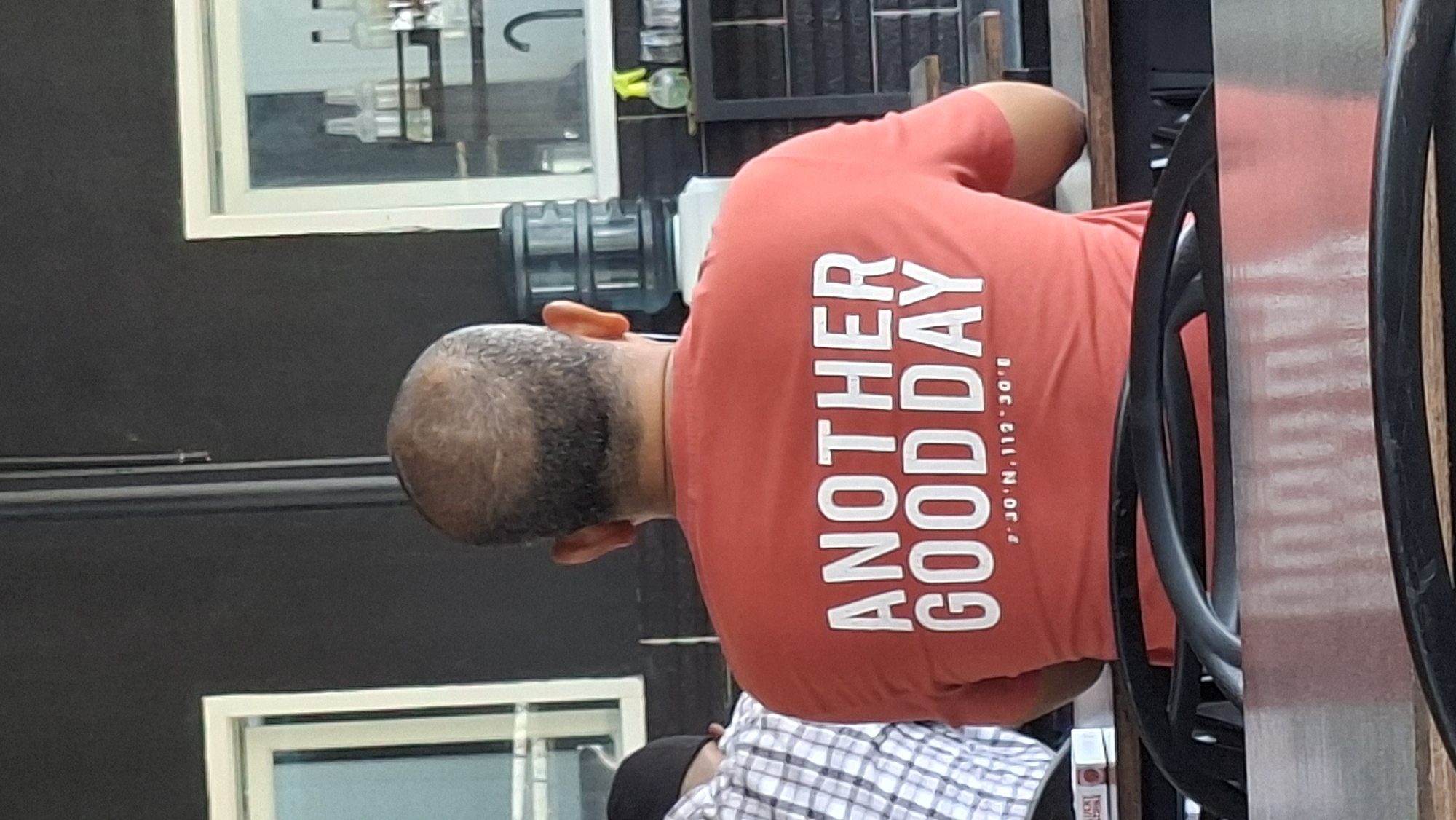The image captures the back of a man wearing a red t-shirt with white text that reads "ANOTHER GOOD DAY" and coordinates below: "2°30'N, 112°30'E". He is seated at a table with a metal surface and black chairs in the foreground, with a dark-colored wall behind him. To his left, another person is visible, wearing a checkered shirt and a black cap. In the background, there are framed windows, shelves with bottles and a dispenser, and a black brick wall. The scene appears to be an indoor setting, possibly a cafe or restaurant. The coordinates indicate a location in Indonesia, potentially within or near Lhokseumawe, Aceh Province.