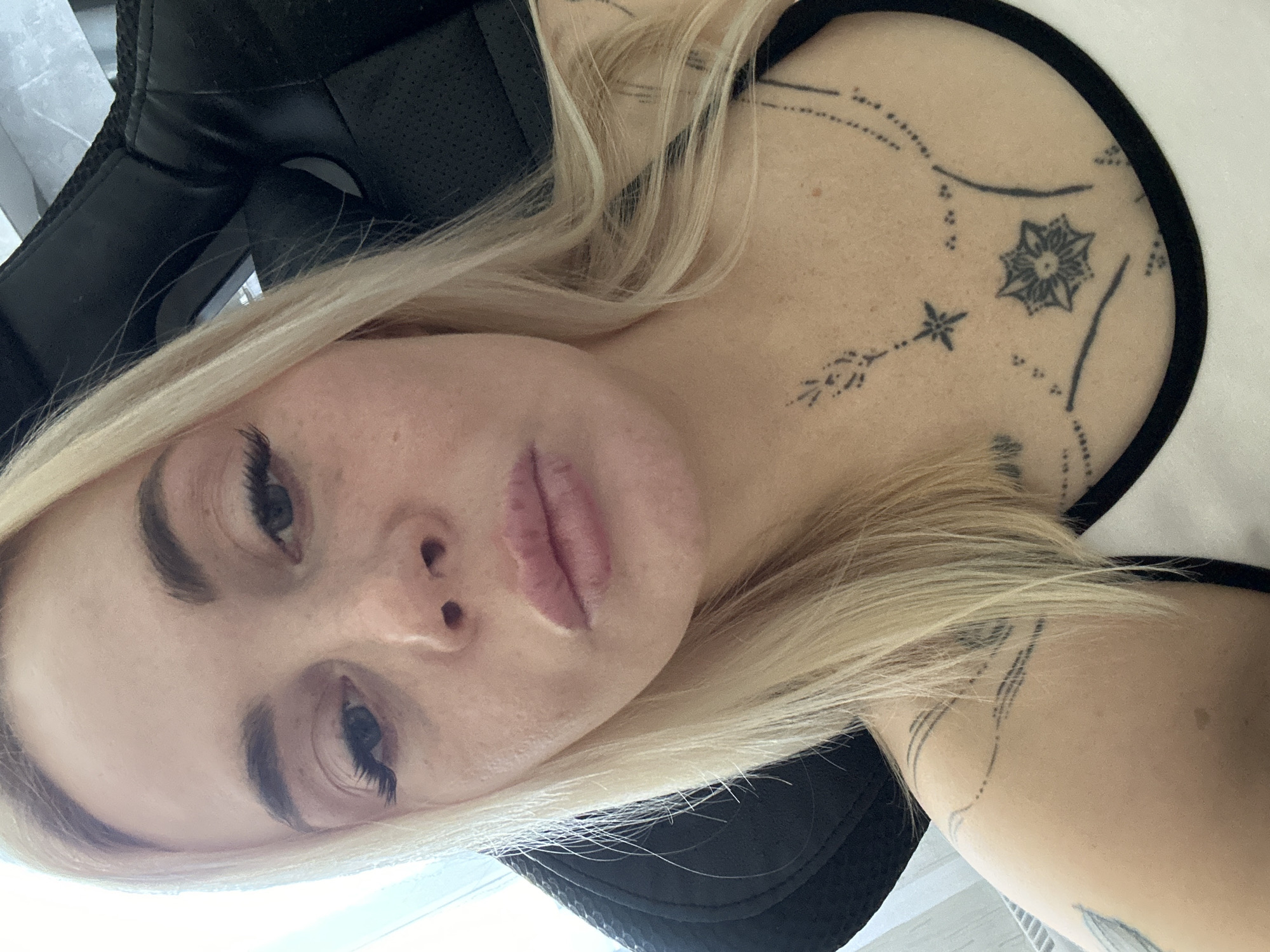 A blonde woman is captured in a close-up, selfie-style shot, looking directly at the camera. She is wearing a white tank top with a black trim, and her chest and shoulders are adorned with various black line tattoos, including a floral-like design on her chest. Her hair is light blonde and falls around her face and shoulders. The background is out of focus, but appears to be an indoor setting with a dark, possibly leather, chair and some light coming from the left. Her skin has a light complexion with some freckles visible on her face.