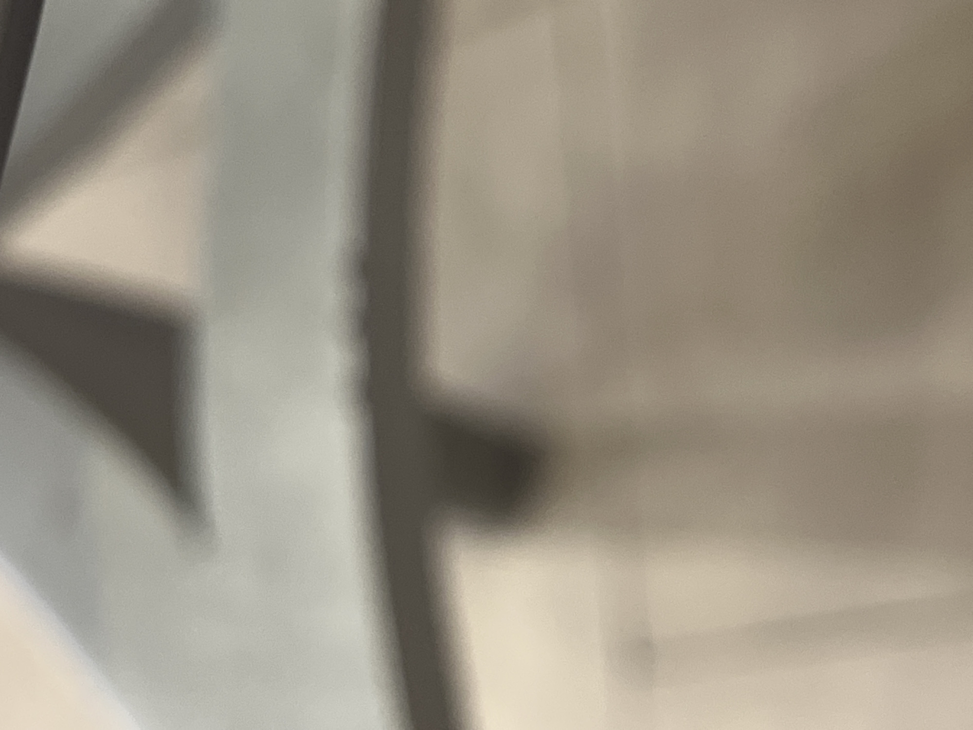 The scene depicts a blurry, abstract interior architectural detail within the city of Casablanca, Morocco. The upper portion of the frame features a dark, horizontal linear element, likely a structural beam or edge. Below this, distinct dark triangular shadows are cast against lighter grey and beige surfaces, suggesting angular forms or reflections from overhead structures. The lower two-thirds of the image comprise a smooth, light beige to brown surface, possibly a floor or wall, exhibiting faint vertical tonal shifts. No individuals, clearly identifiable objects, or specific actions are discernible within the frame.